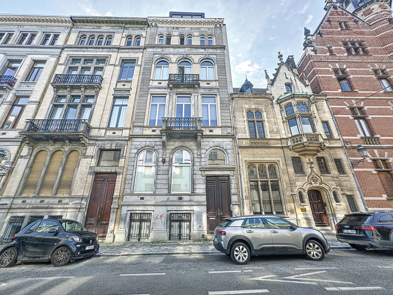 Wijze foto 10 Een rij historische stenen herenhuizen met sierlijke balkons aan een geplaveide straat in de stad, met geparkeerde auto’s langs de stoeprand.
