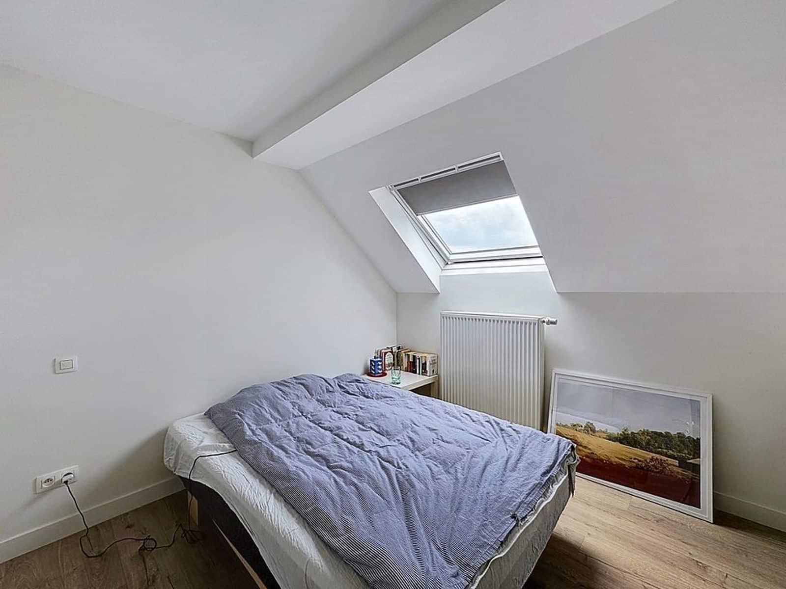 Chambre Mansardée Lumineuse – You Real Estate Agency Chambre mansardée lumineuse avec lit, parquet et fenêtre de toit en appartement.