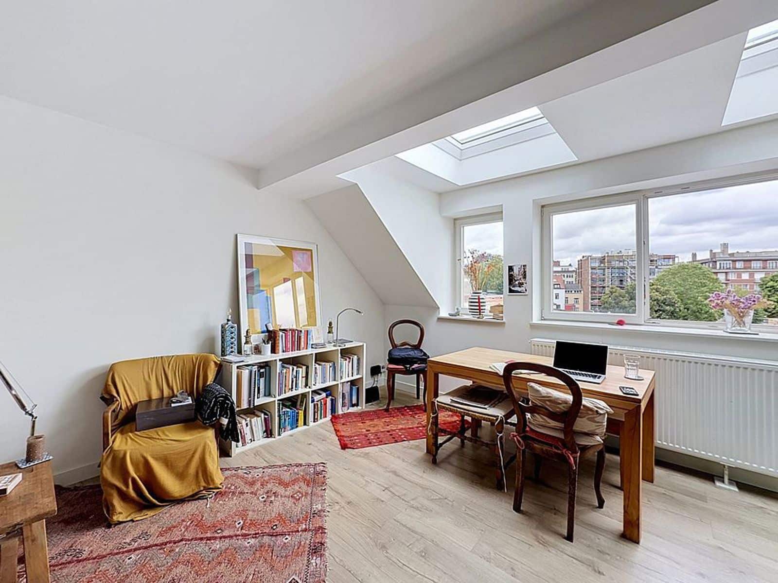whise photo 2 Bright attic home office with skylights, a wooden desk and laptop facing a large window overlooking the city, plus a bookshelf and chairs nearby.