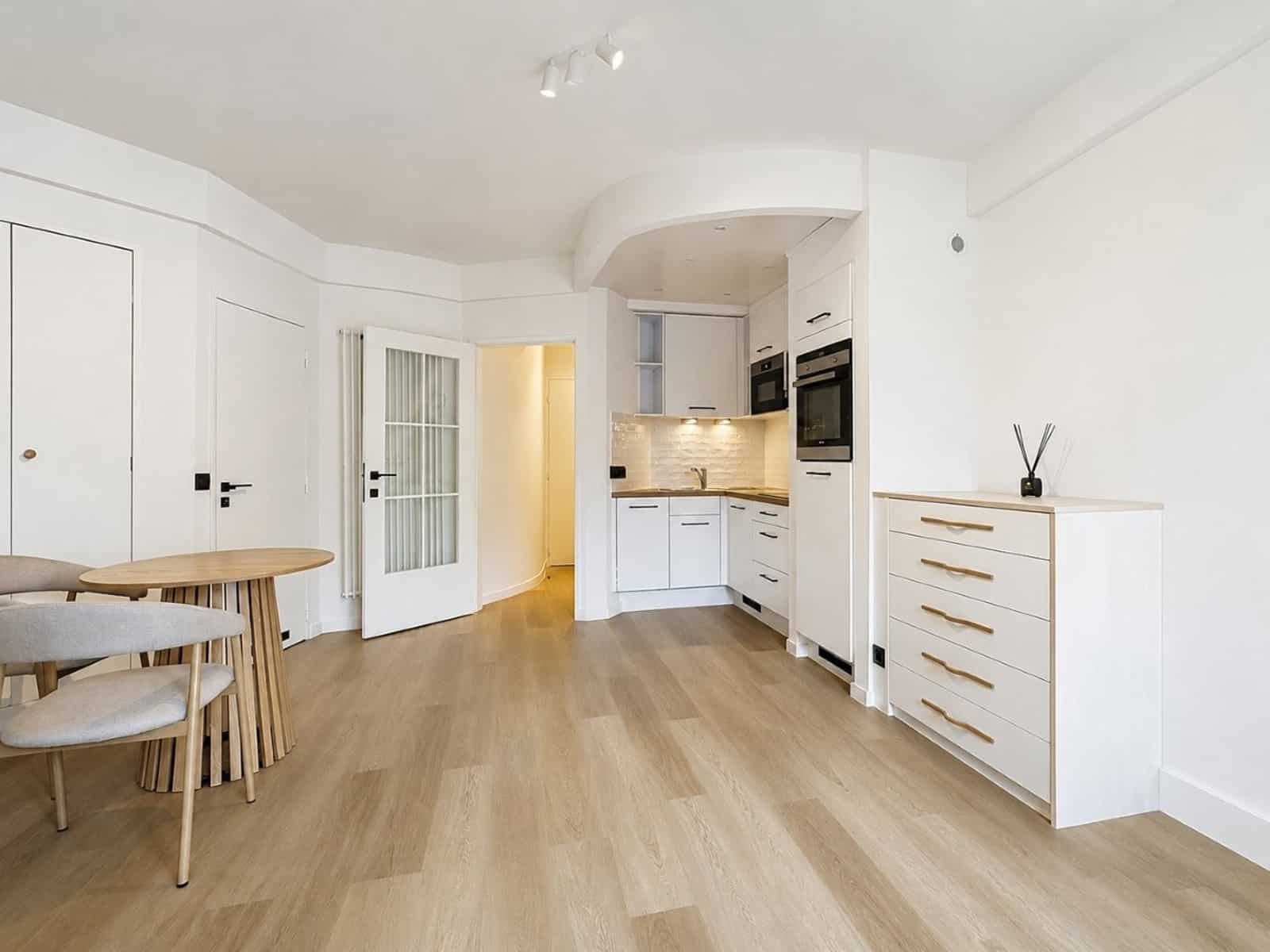 whise photo 7 Bright open-plan kitchen with white cabinets, built-in oven, and light wood flooring connected to a dining area with a round wooden table.