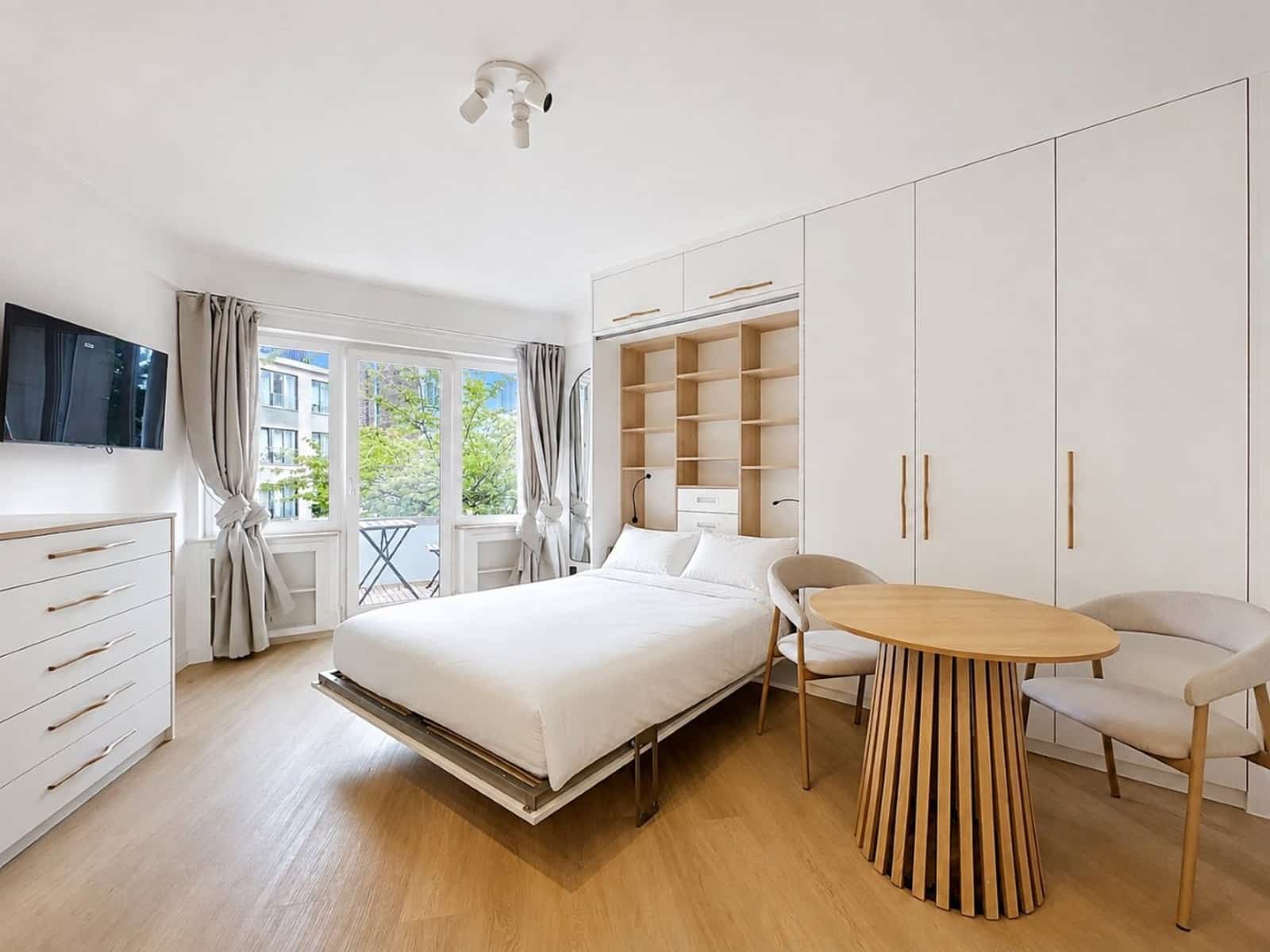 whise photo 2 Bright modern bedroom with white built-in wardrobes, a low bed, round wooden table and chairs, and a balcony door with curtains.
