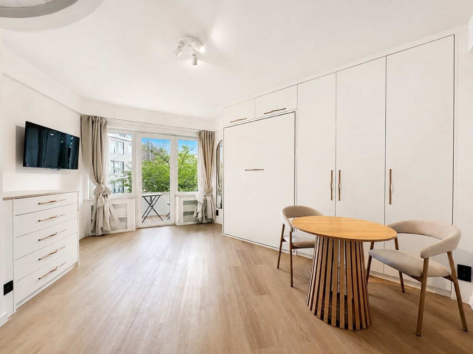 whise photo 6 Bright modern living space with white built-in wardrobes, a round wooden table and two beige chairs, and a wall-mounted TV. Balcony doors and greenery visible outside.