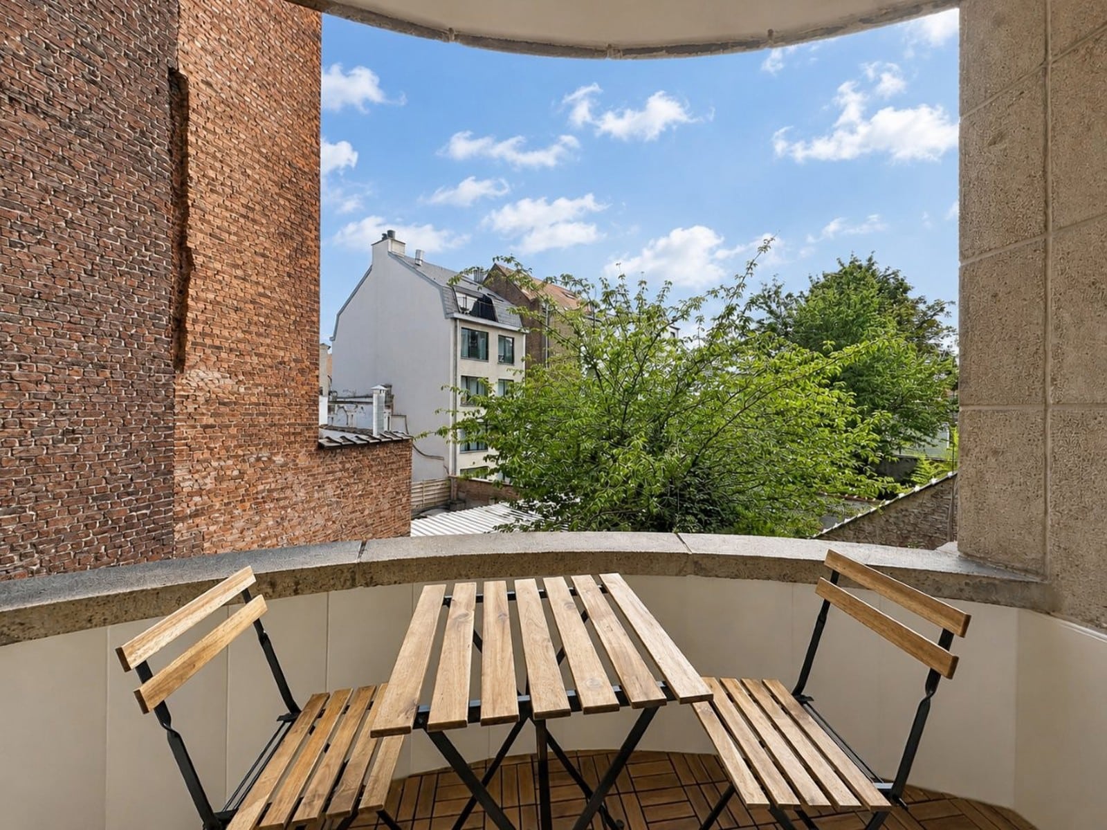 whise photo 8 Cozy balcony with a wooden table and chairs, brick wall on the left and a green tree against a blue sky backdrop.
