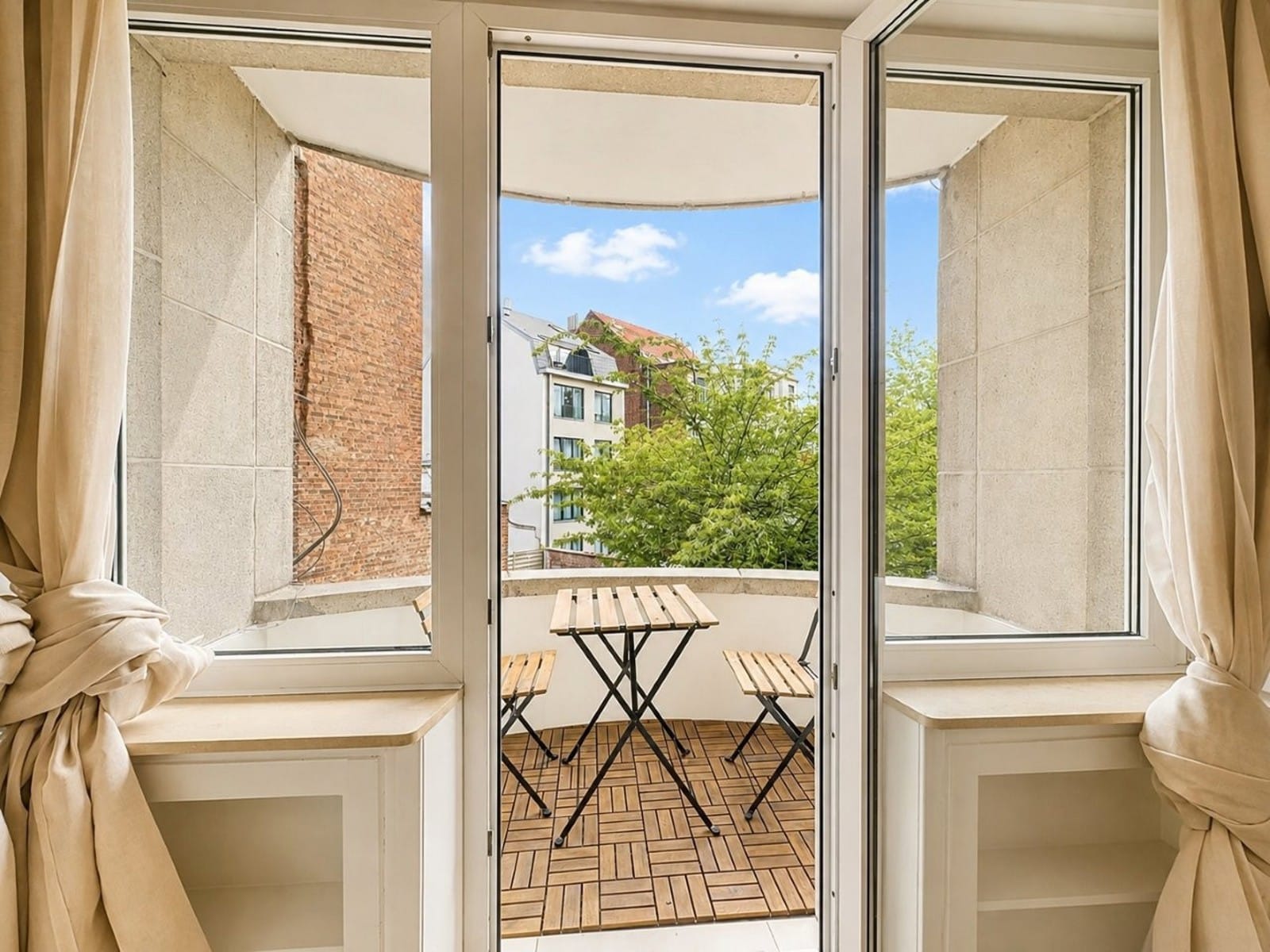 whise photo 9 Open French doors to a small balcony with a wooden bistro table and two chairs, brick buildings and greenery beyond.