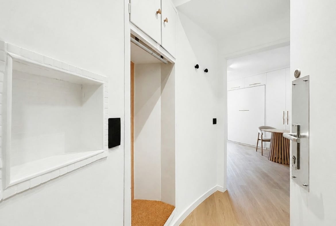 Bright white hallway with an open elevator alcove on the left and a curved wood floor leading to a room with a round table.