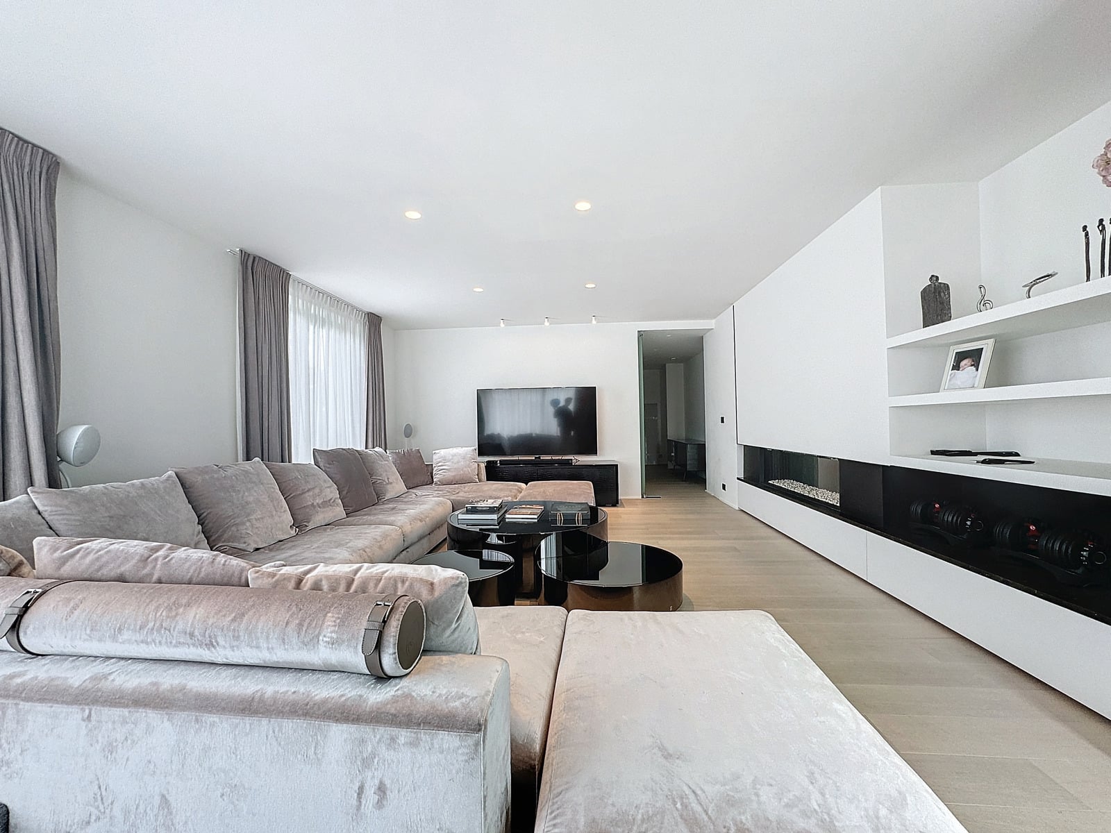 whise photo 4 Bright modern living room with a large light-gray sectional, multiple black glass coffee tables, and a wall-mounted TV. A white built-in cabinet runs along the right wall.