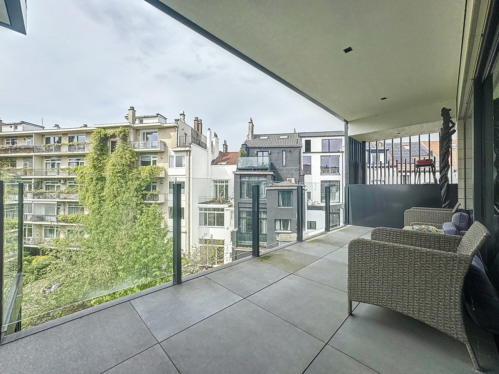 whise photo 14 Modern balcony with glass railing and gray tile floor, outdoor wicker seating facing a row of urban buildings and trees.