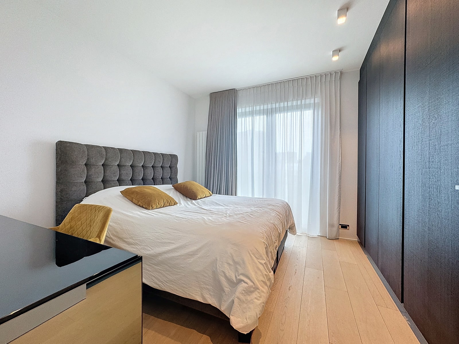 whise photo 19 Modern bedroom with a gray tufted headboard, white bedding, and two mustard pillows; large window with sheer white curtains and dark wardrobe along the right wall.