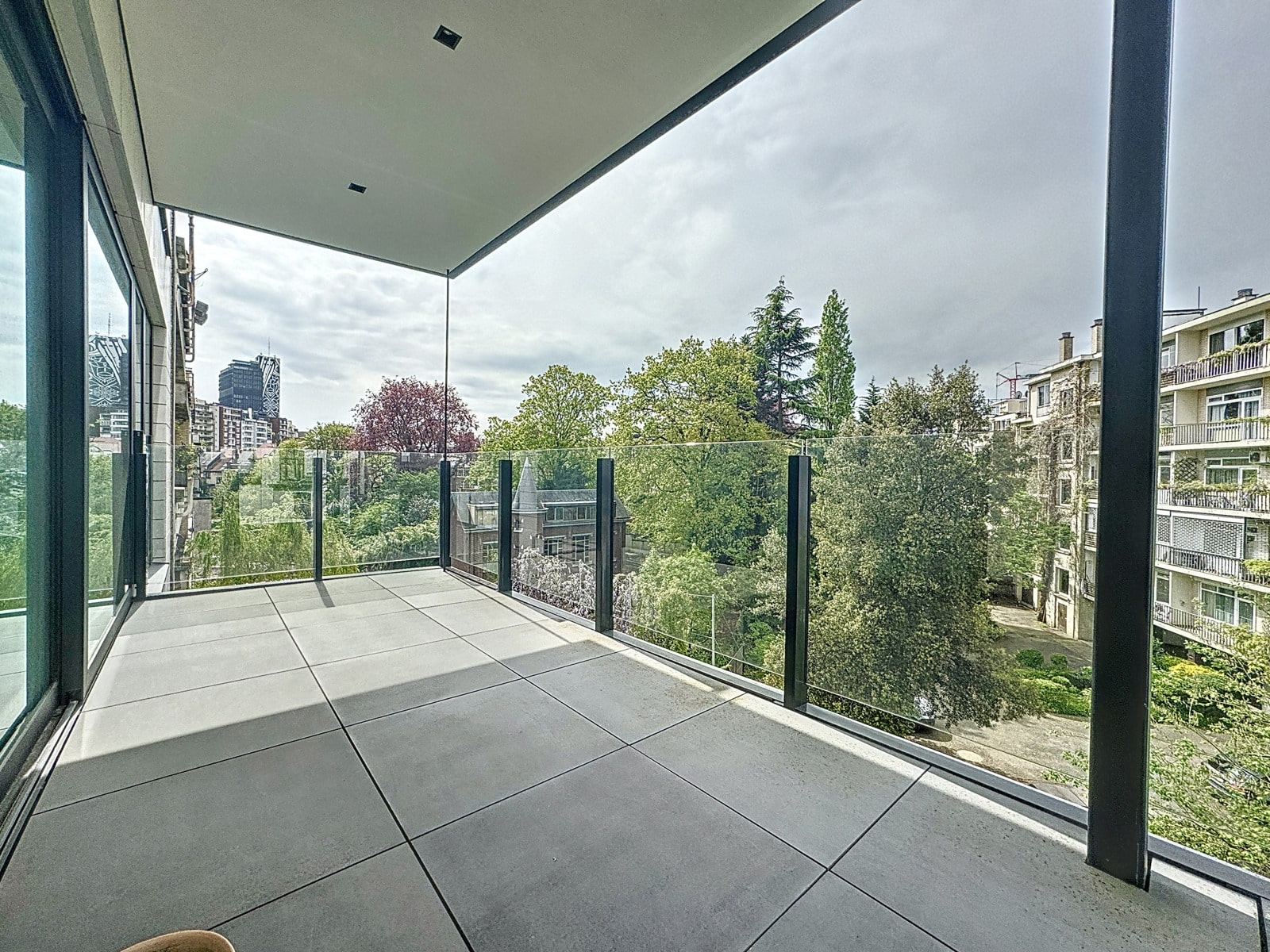 whise photo 8 Modern balcony with a glass railing overlooking a leafy cityscape and neighboring buildings.