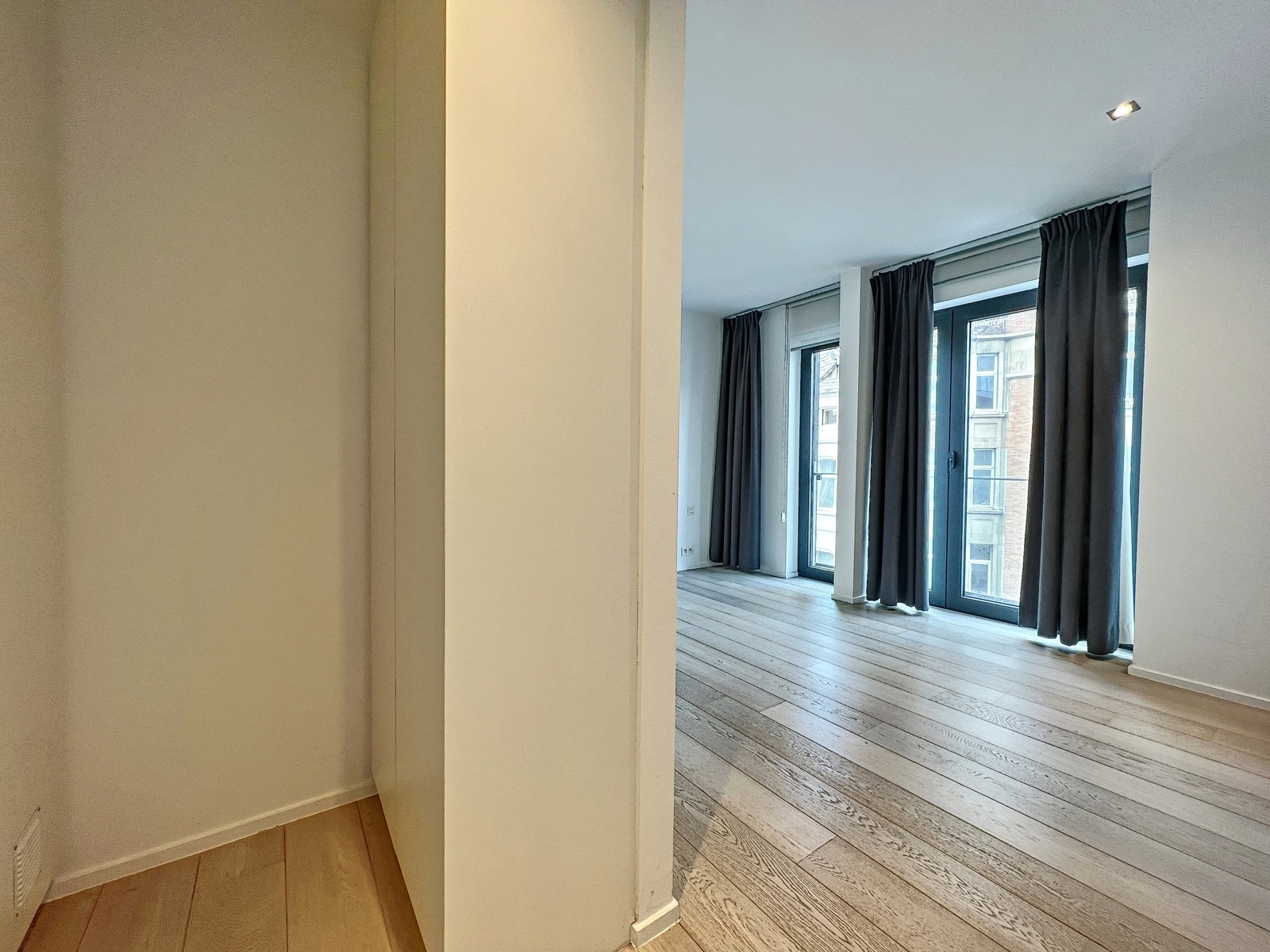 whise photo 9 Bright living area with large floor-to-ceiling windows and dark curtains, light wood floors, white walls, and a partial wall divider.