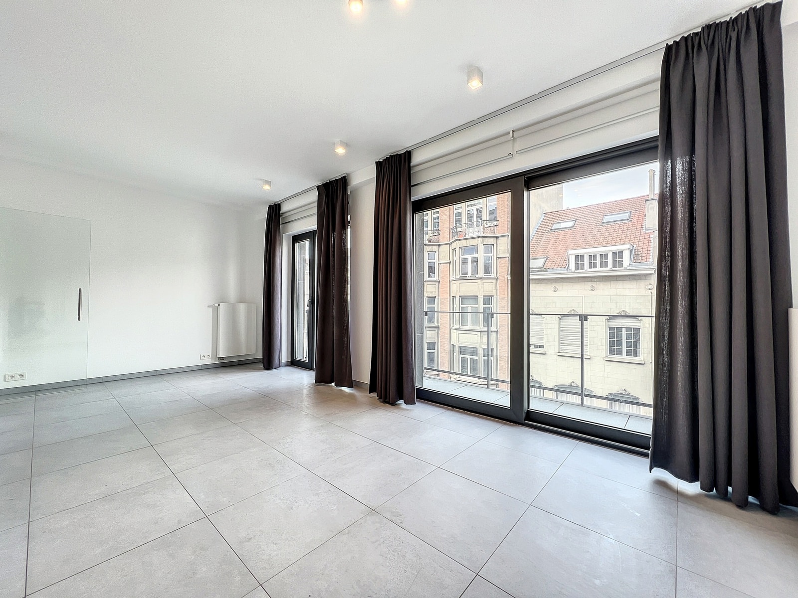 whise photo 2 Bright empty living room with large floor-to-ceiling windows and dark drapes.