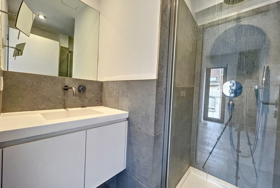 White bathroom vanity with a wide sink, grey tiled walls, and a glass shower enclosure with a rainfall shower head on the ceiling and a handheld wand.