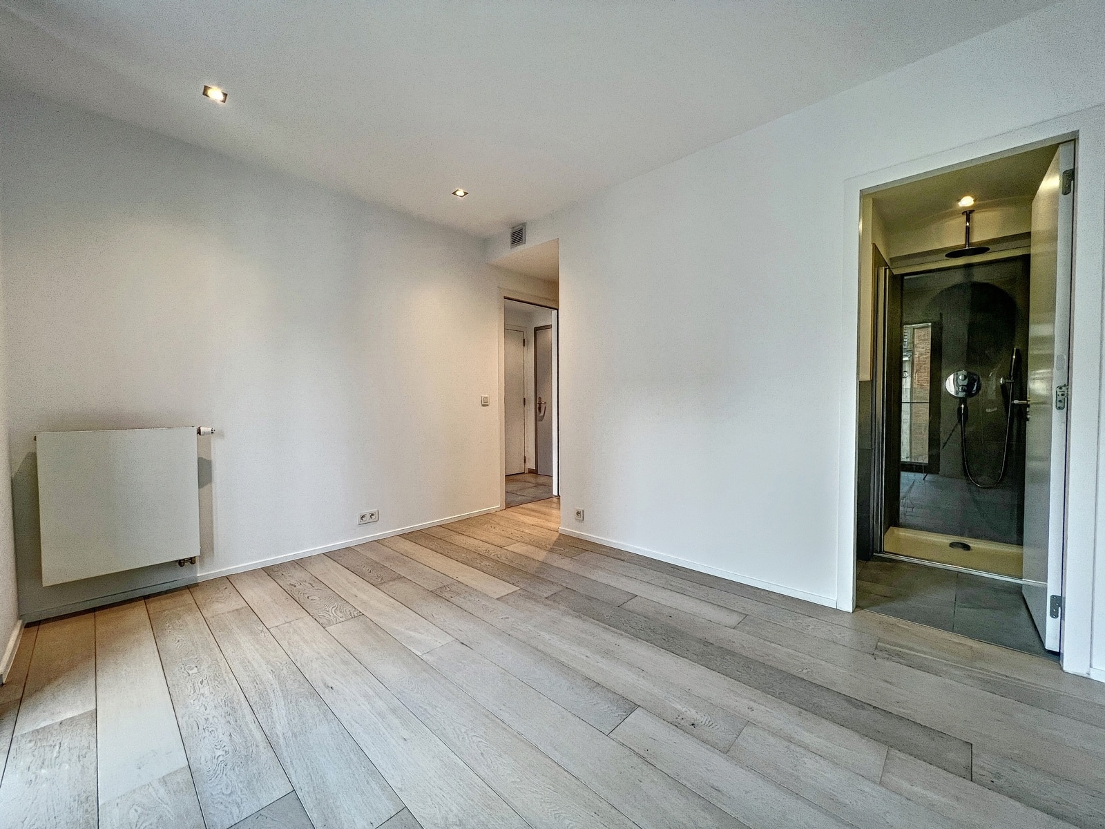 whise photo 12 Empty modern room with light wood flooring, white walls, a wall-mounted radiator on the left, and a doorway to a hallway; a glass-enclosed shower is visible on the right side.