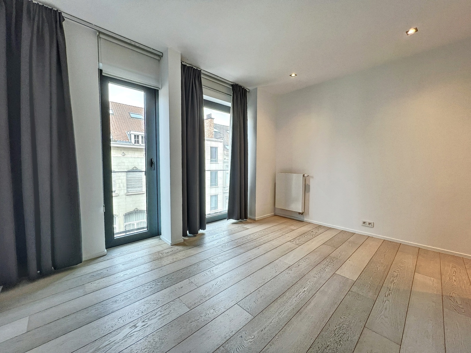 whise photo 11 Bright empty room with two tall windows/doors, dark gray curtains, and light gray wooden flooring; a wall-mounted radiator is on the far wall.