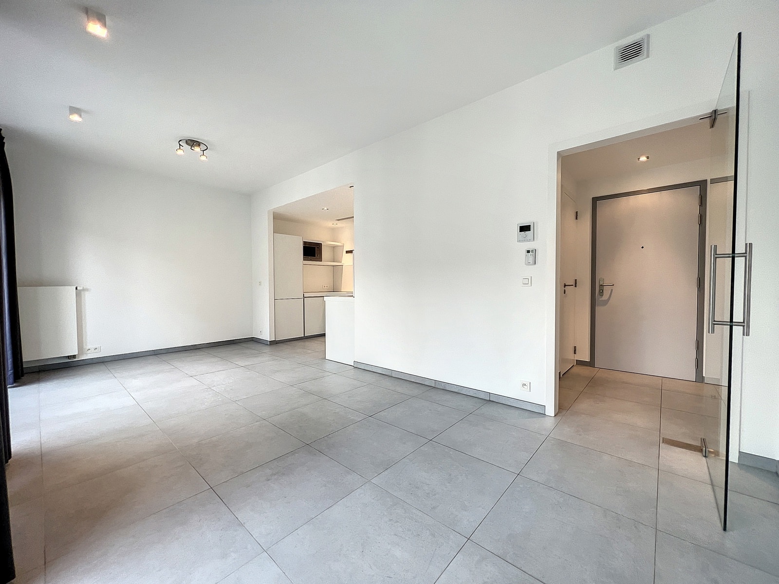 whise photo 6 Empty living room with large gray tile floor, white walls, and an open kitchen to the left; glass door on the right.