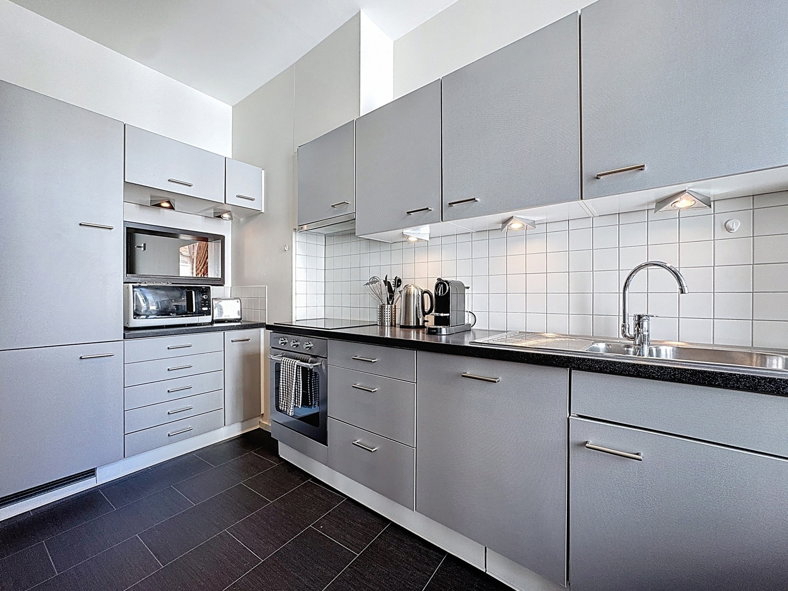 Cuisine Moderne Équipée – You Real Estate Agency Modern grey kitchen with upper and lower cabinets, white tiled backsplash, and a stainless steel sink with a glossy countertop.