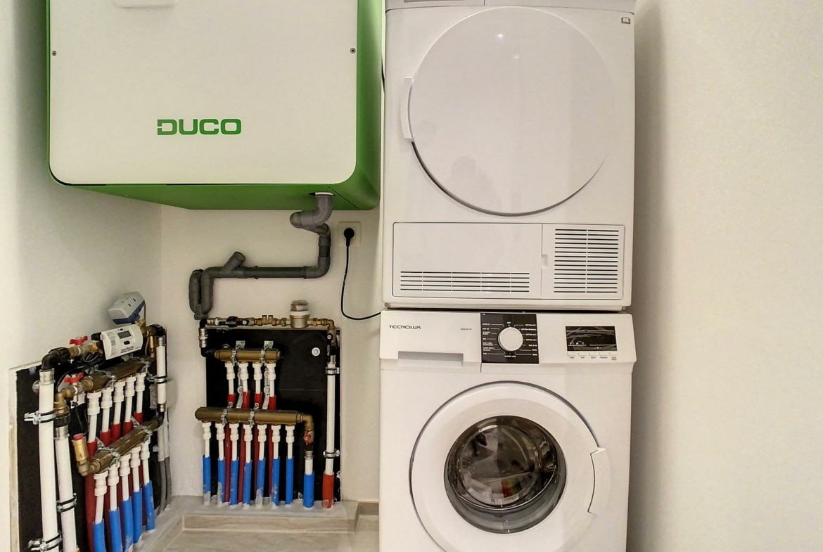 Rechts een gestapelde witte wasmachine en droger, links een groene Duco-kast en daaronder zichtbare leidingen.