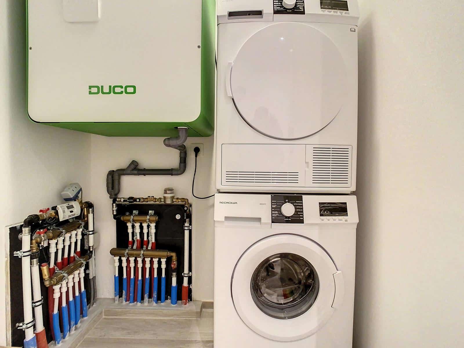 Moderne, volledig uitgeruste wasruimte – You Real Estate Agency Rechts een gestapelde witte wasmachine en droger, links een groene Duco-kast en daaronder zichtbare leidingen.
