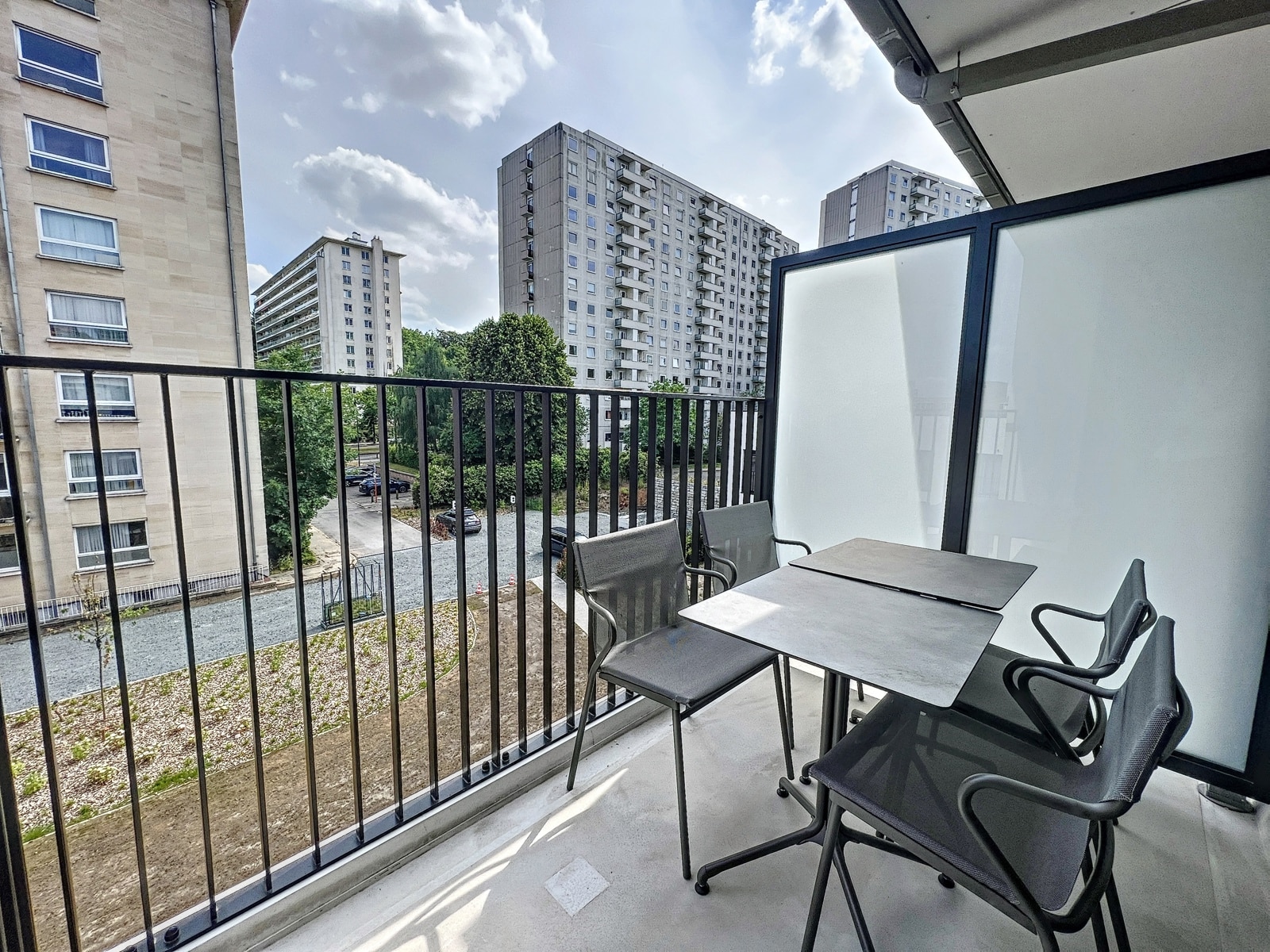 Modern balkon met uitzicht op de woonwijk – You Real Estate Agency Modern appartementsterras met tuinmeubilair en uitzicht op een met bomen omgeven wooncomplex.