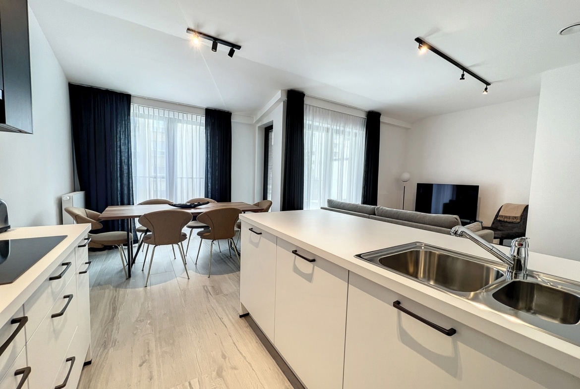 Een open keuken met eethoek, voorzien van een wit kookeiland, een spoelbak en een houten eettafel met beige stoelen; een woonkamer met op de achtergrond een tv en grote ramen met donkere gordijnen.