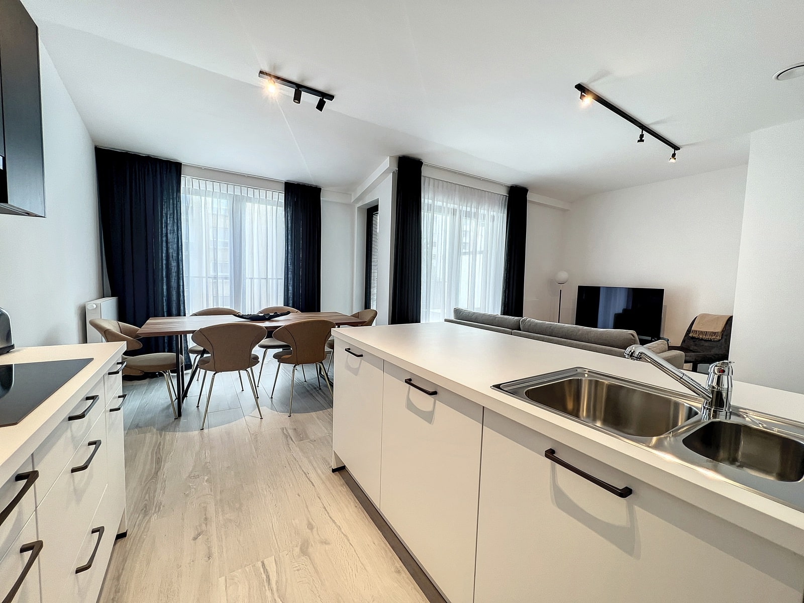 foto 5 Een open keuken met eethoek, voorzien van een wit kookeiland, een spoelbak en een houten eettafel met beige stoelen; een woonkamer met op de achtergrond een tv en grote ramen met donkere gordijnen.