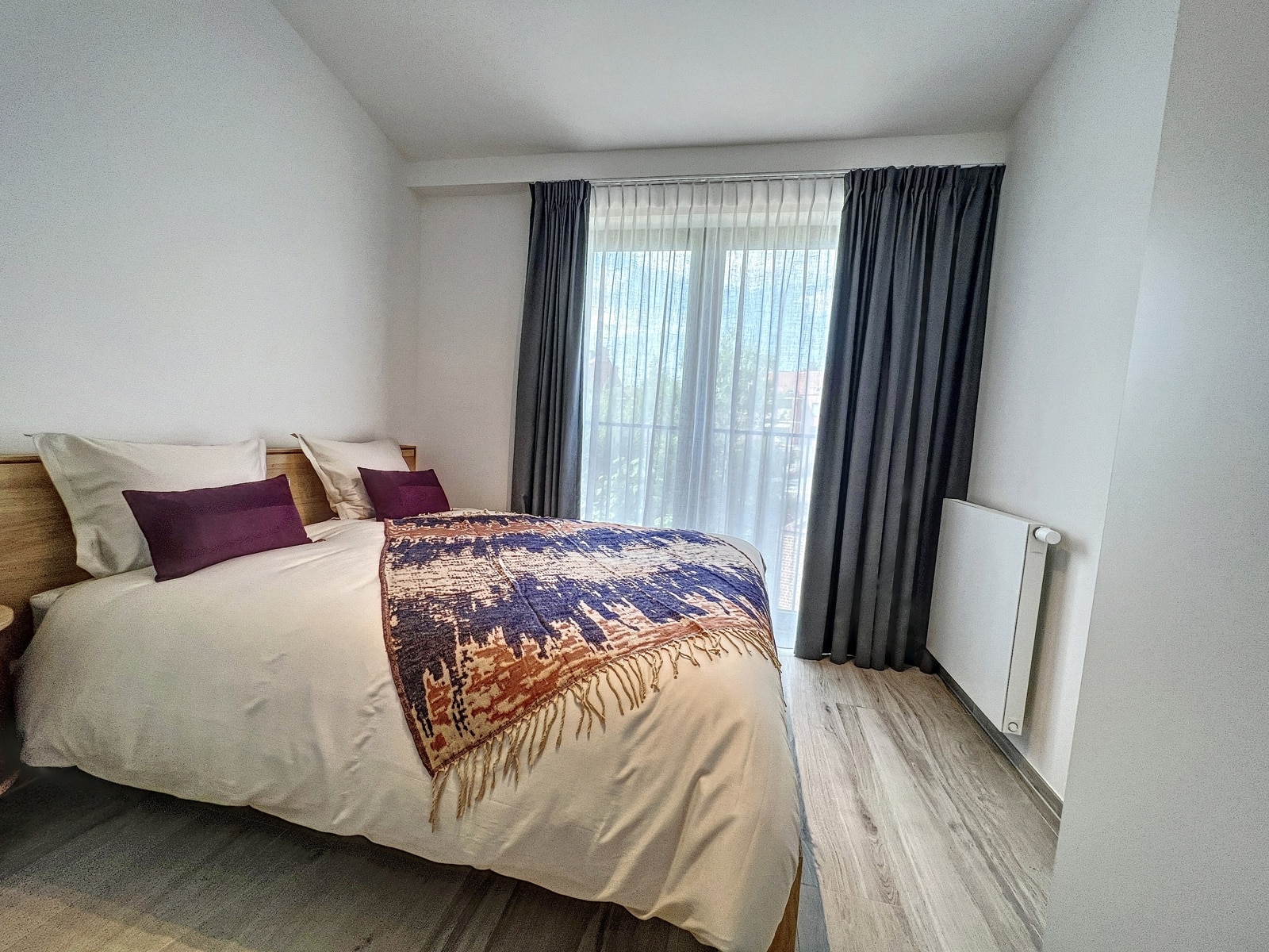 Lichte kamer met tweepersoonsbed - U Vastgoedkantoor Een queensize bed met een wit dekbed, paarse kussens en een plaid met patroon in een lichte slaapkamer bij een groot raam met vitrages en grijze gordijnen.