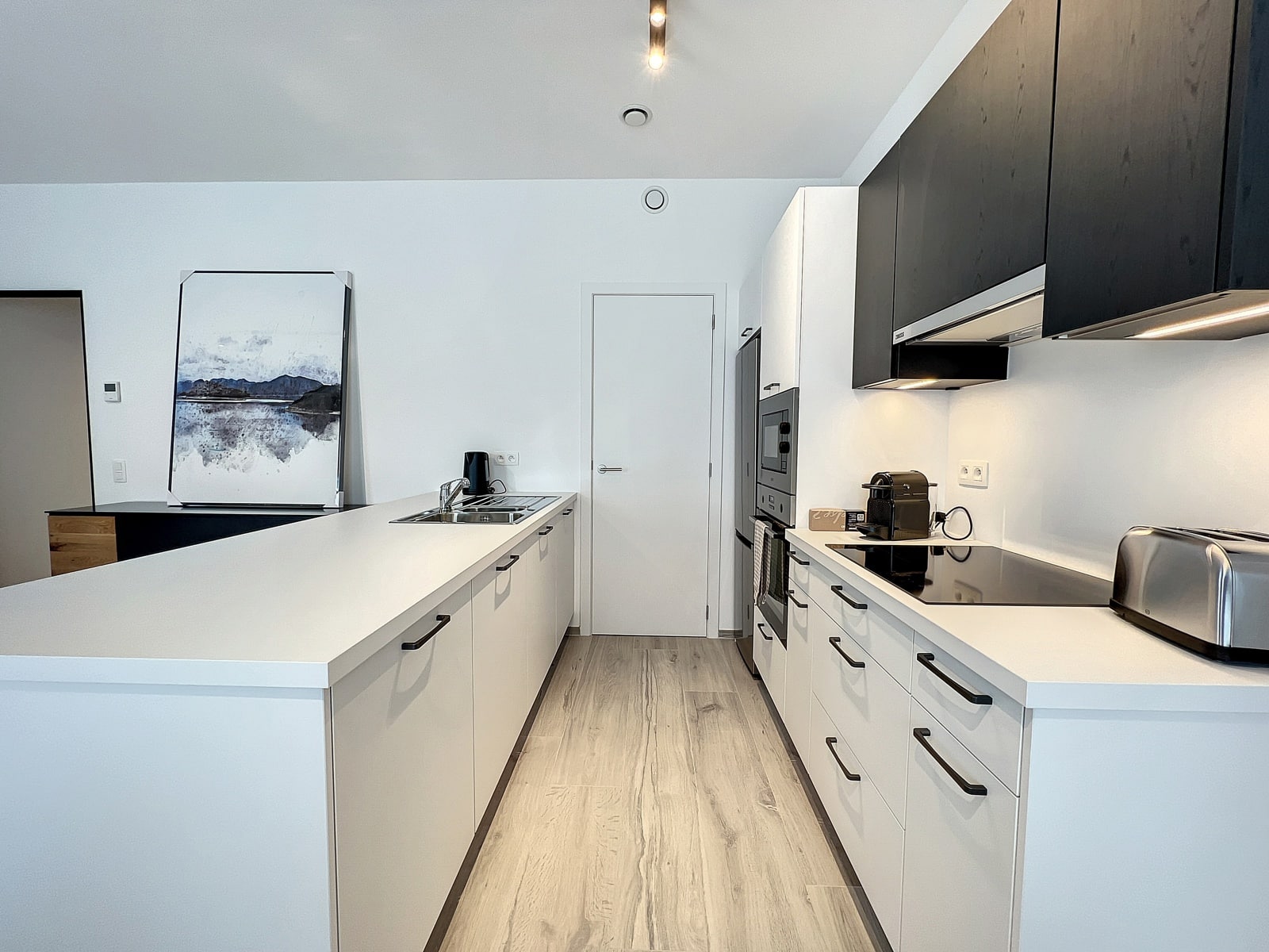 foto 4 Moderne, lichte keuken met witte kasten en een lang kookeiland, daarboven zwarte kastdeuren en een ingelijst landschapsschilderij dat tegen de muur leunt.