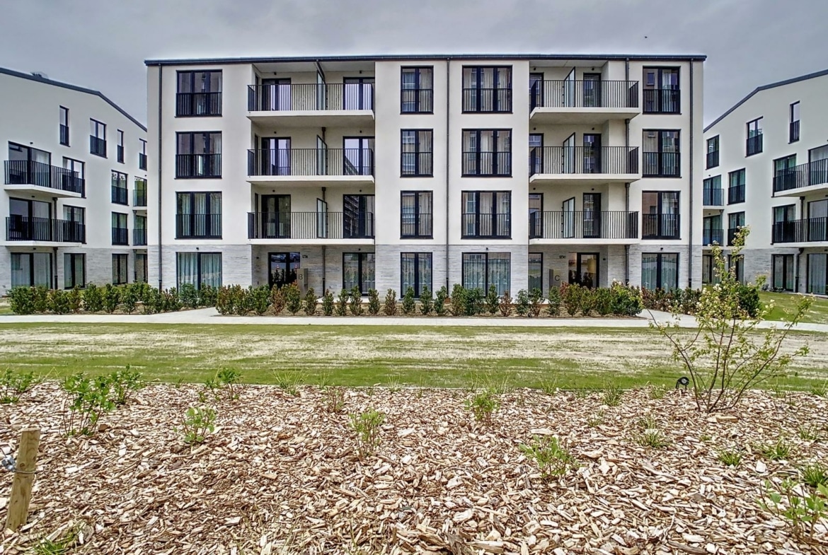 Modern wit appartementencomplex met drie verdiepingen en balkons, gelegen rondom een aangelegde binnenplaats met struiken en mulch.