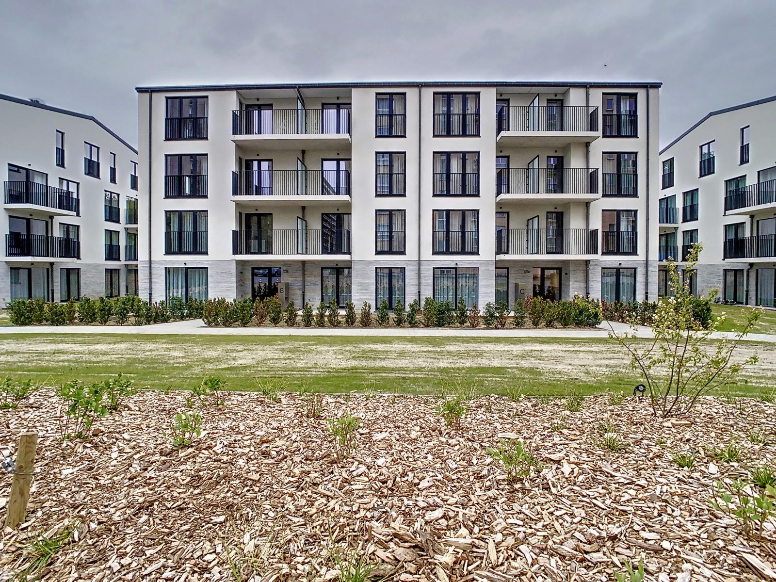 Wijze foto 14 Modern wit appartementencomplex met drie verdiepingen en balkons, gelegen rondom een aangelegde binnenplaats met struiken en mulch.