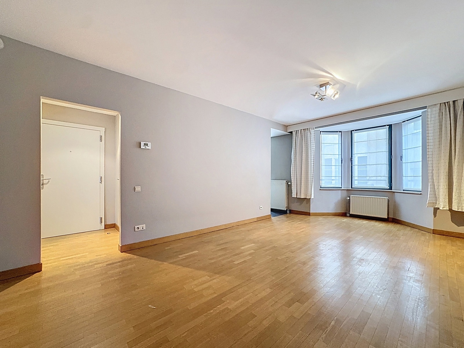 Lichte woonkamer met parketvloer - You Real Estate Agency Lichte, lege woonkamer met parketvloer, grote ramen en radiatoren in een modern appartement.