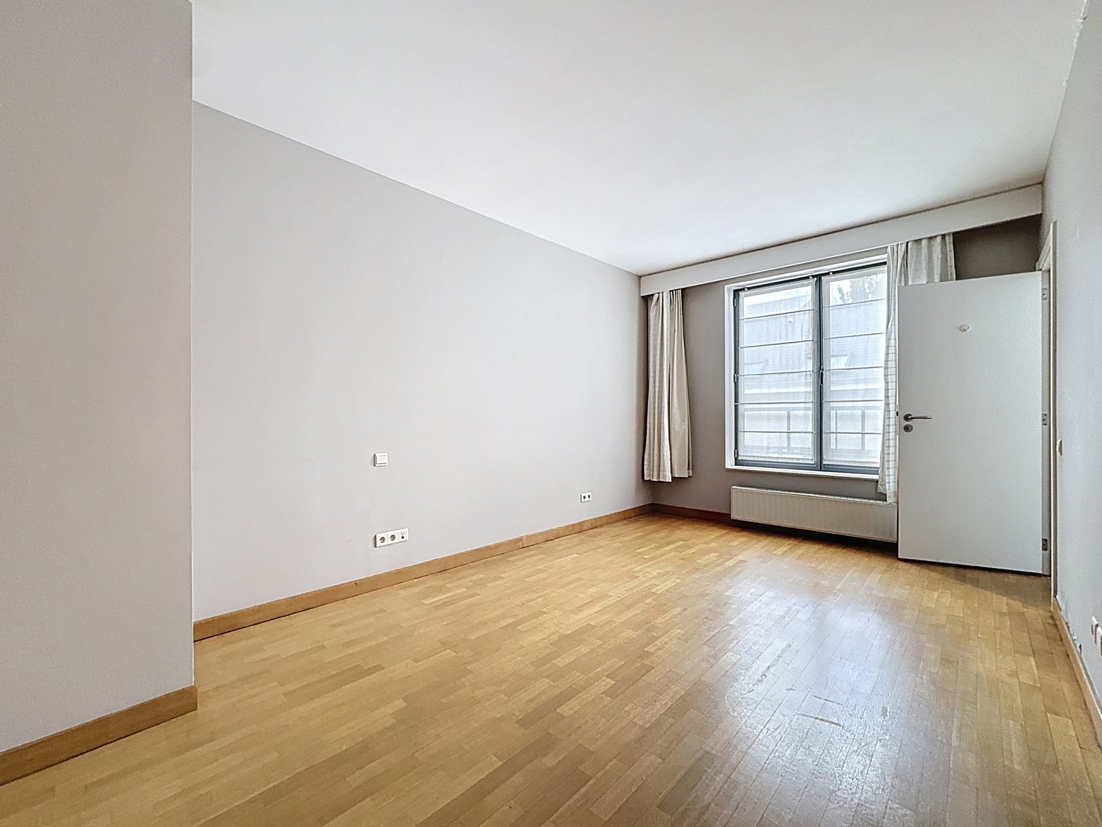 Lichte woning met parketvloer – You Real Estate Agency Lichte woonkamer met parketvloer, raam, gordijnen en radiator.