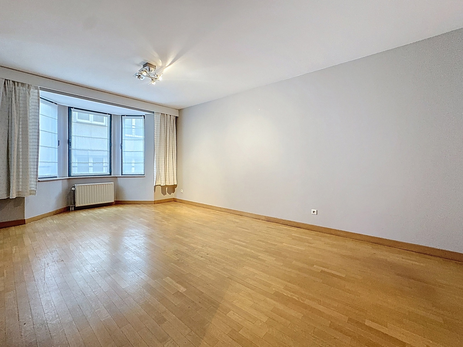 Lichte woonkamer met parketvloer – You Real Estate Agency Lichte, lege woonkamer met parketvloer, grote ramen en een radiator in het appartement.