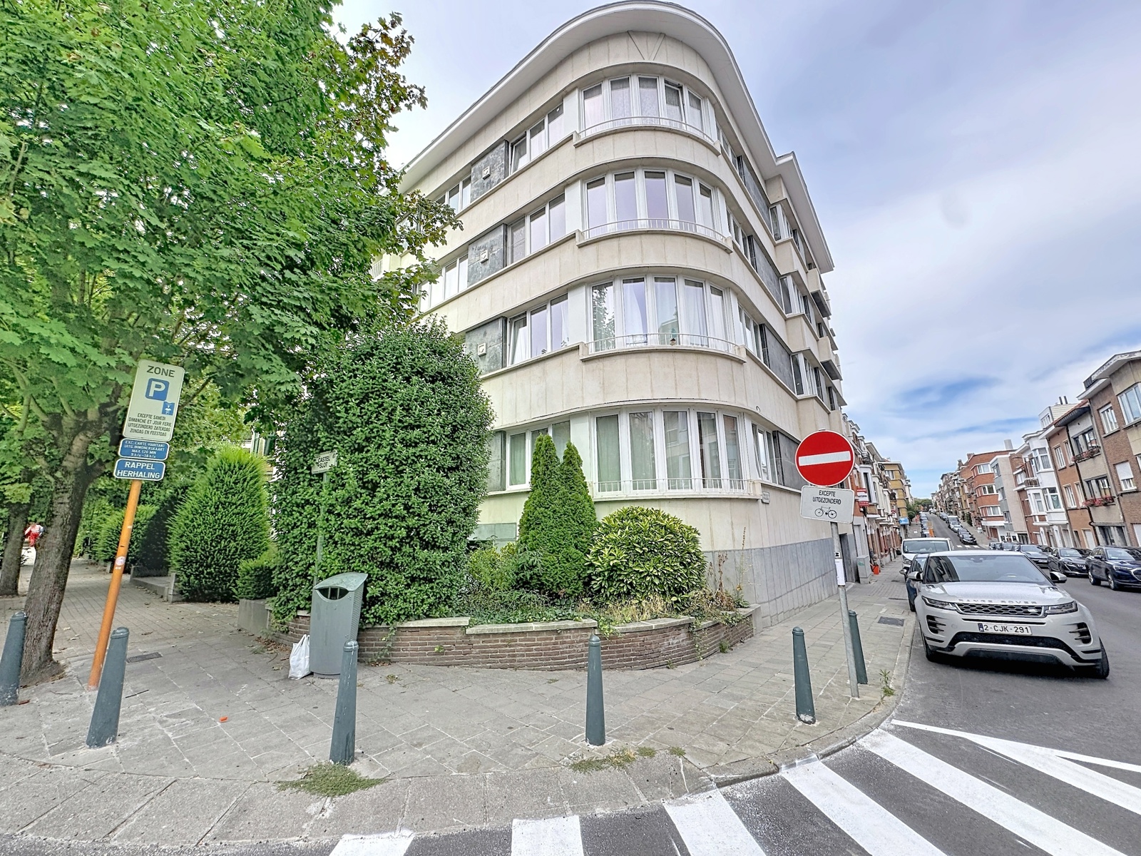 Façade d’Immeuble d’Angle – You Real Estate Agency Immeuble d’appartements moderne en angle avec rue résidentielle arborée.