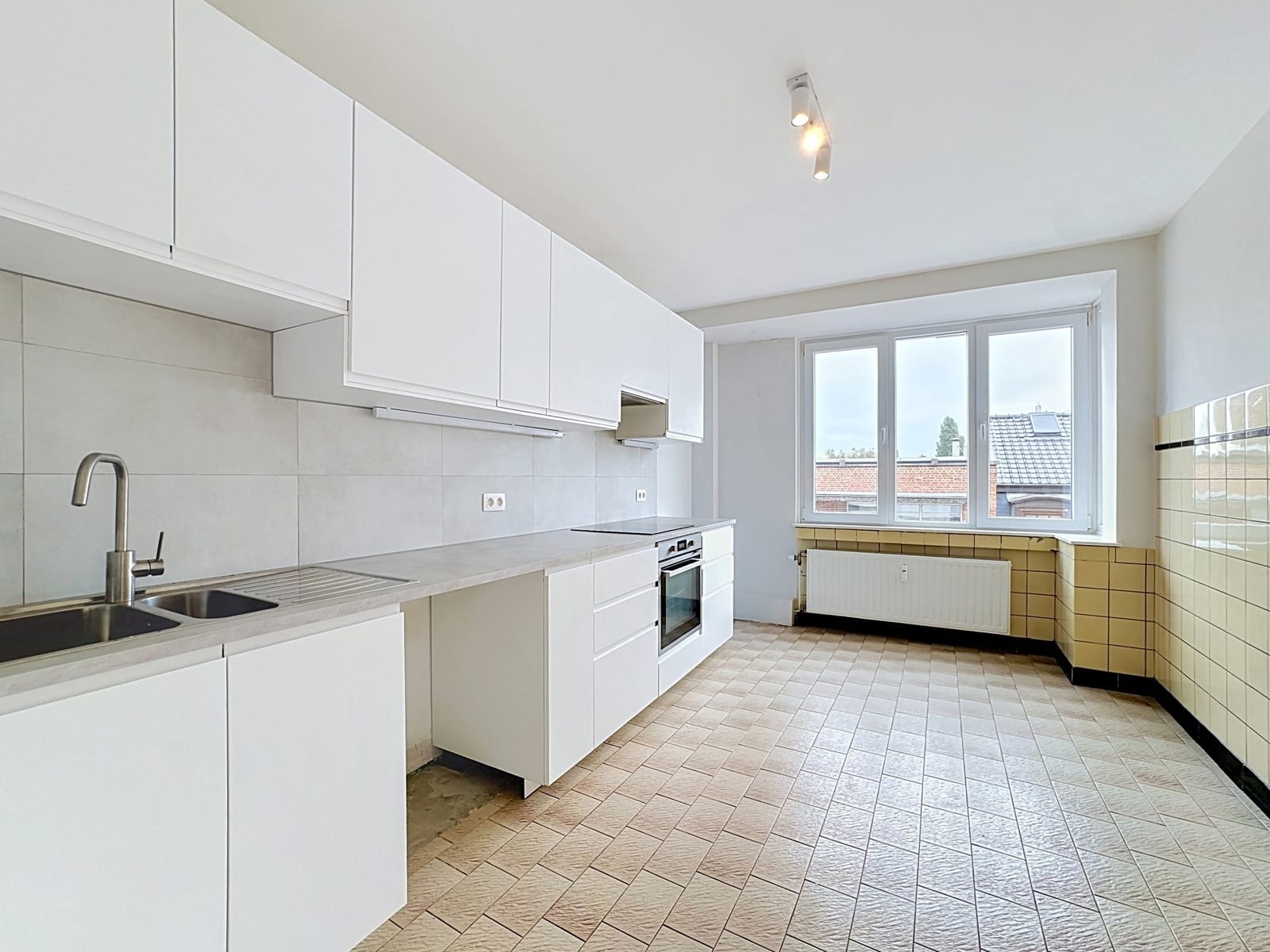 Cuisine Équipée Lumineuse – You Real Estate Agency Cuisine équipée lumineuse d’appartement avec fenêtre, rangements blancs et carrelage beige.