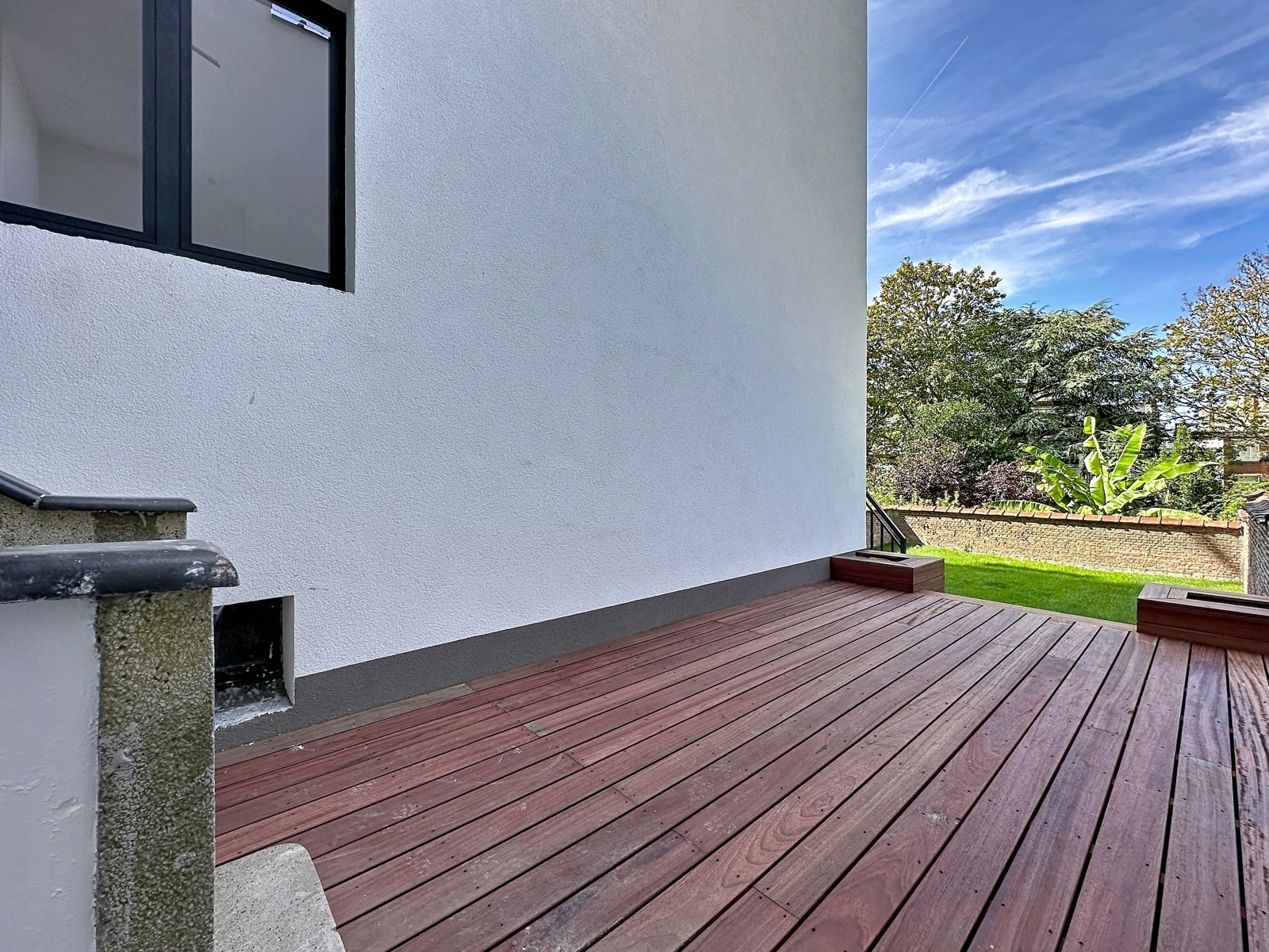Terrasse en Bois et Jardin Verdoyant – You Real Estate Agency Wooden deck attached to a white stucco wall, with steps and a grassy yard beyond under a blue sky.