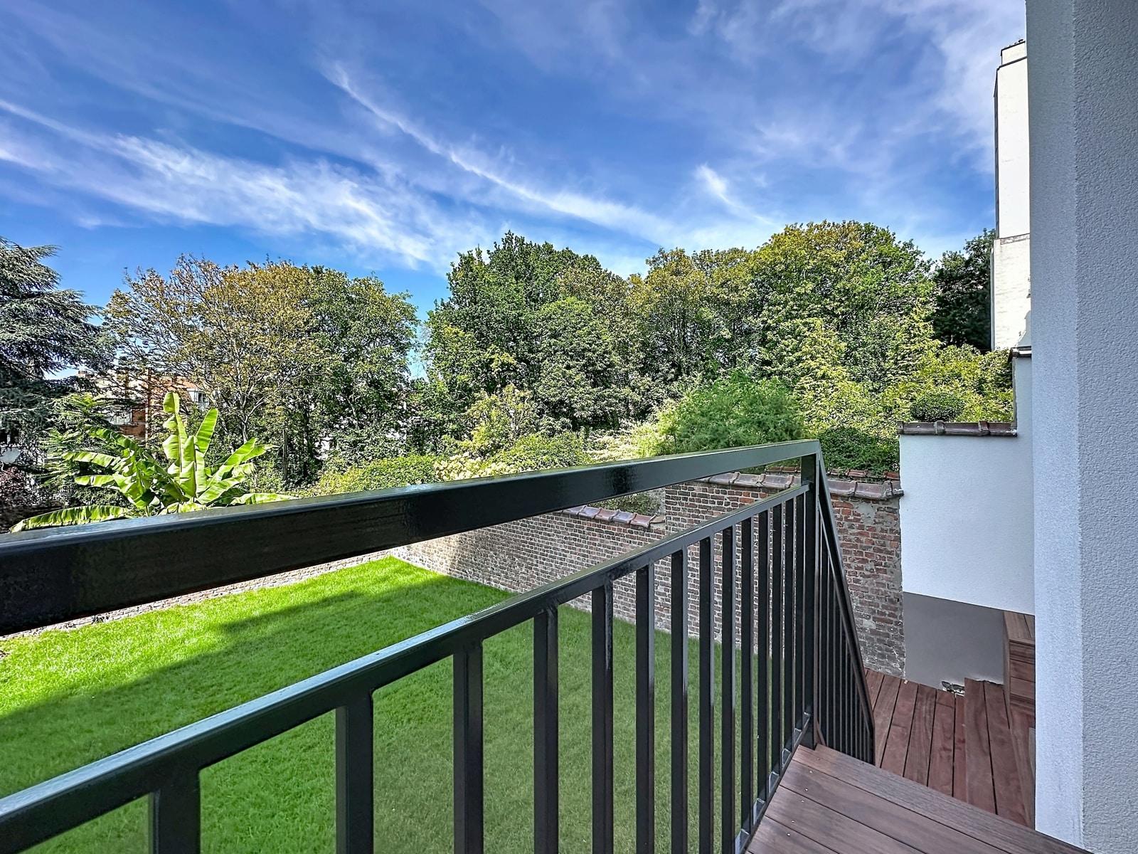 Terrasse Moderne Avec Vue Sur Jardin – You Real Estate Agency View from a balcony: dark metal railing and wooden deck overlook a green lawn, brick wall, and dense trees under a blue sky with wispy clouds.