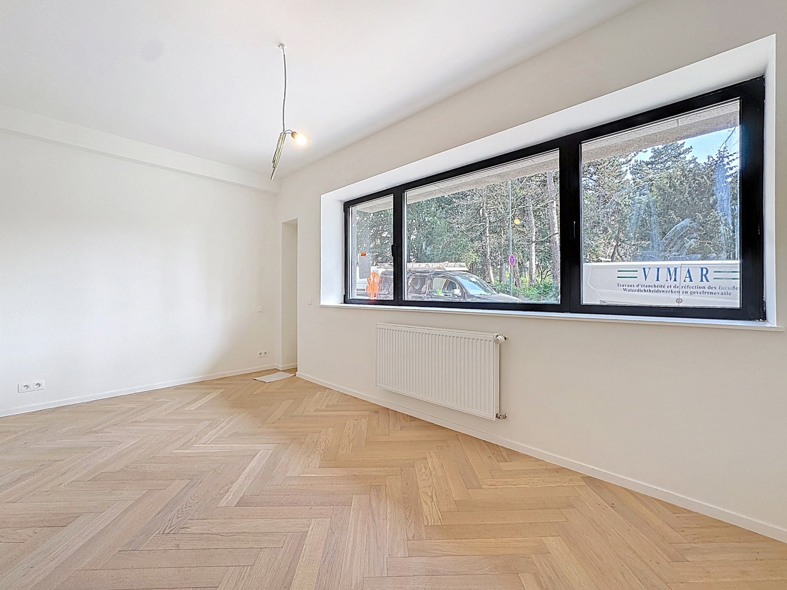whise photo 22 Empty white room with herringbone wooden floor, large black-framed horizontal window, radiator beneath, and a single hanging bare bulb; outside view shows trees and a parked car.