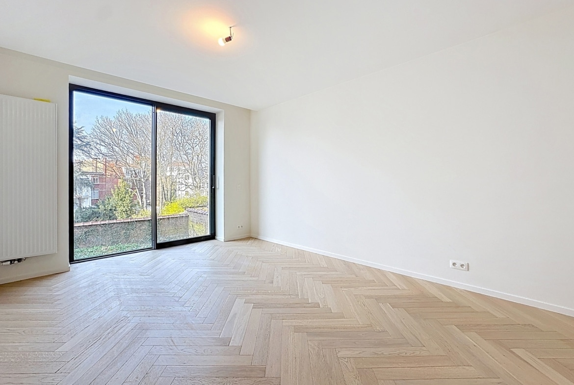 Bright, empty room with light wood herringbone flooring, white walls, and a large sliding glass door opening to an outdoors view.