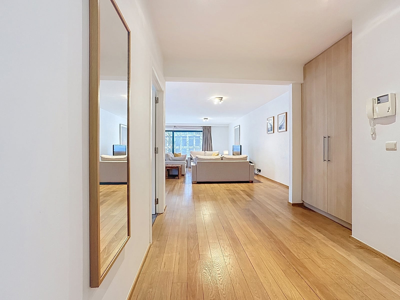 whise photo 3 Hallway leading to a bright living room with beige sofas, a wooden coffee table, and a large window.