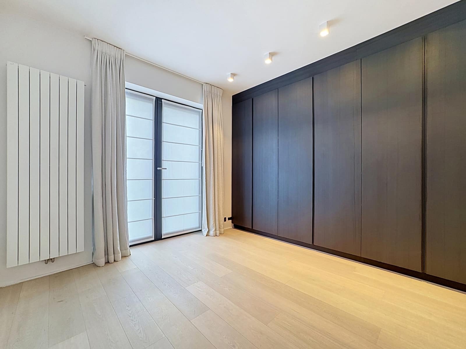 Modern Bedroom with Built-in Walk-In Closet – You Real Estate Agency A modern apartment bedroom with a built-in walk-in closet, light-colored hardwood floors, and a large picture window.