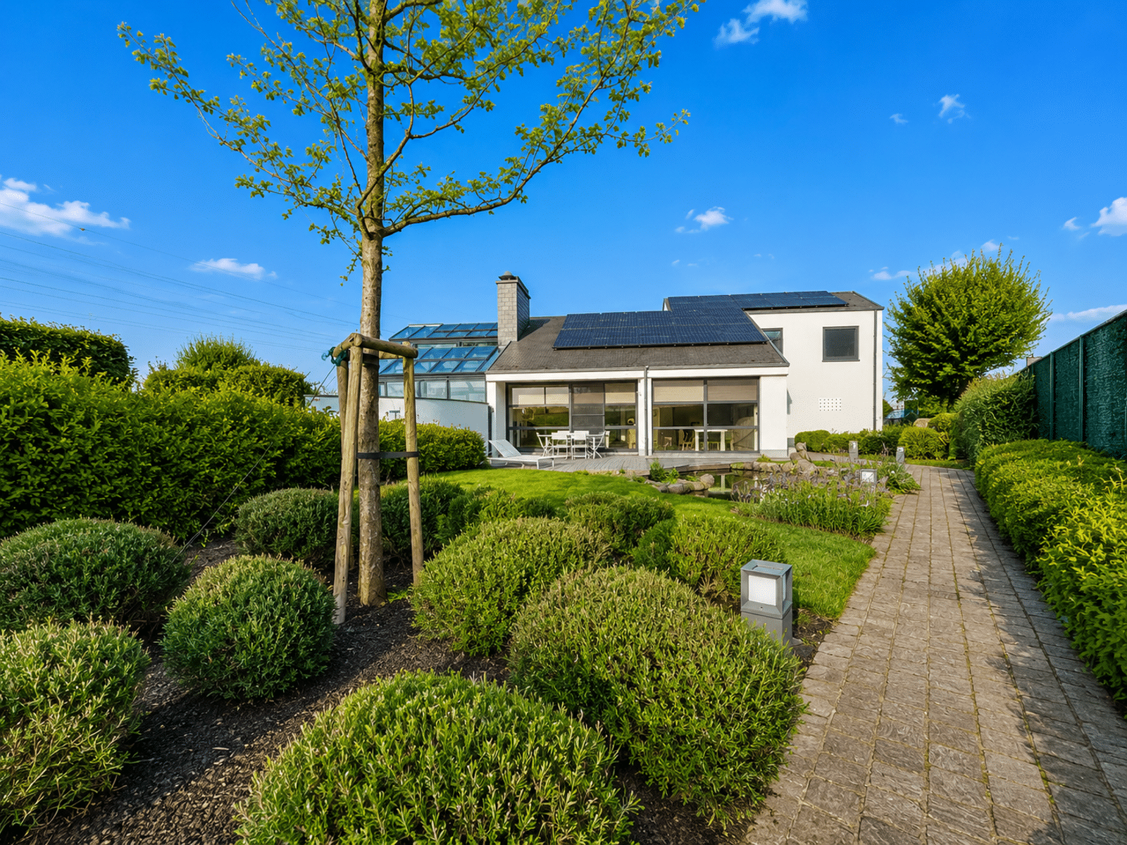 whise photo 3 A modern white house with rooftop solar panels, a glass addition, and a well-kept garden with a stone path under a blue sky.