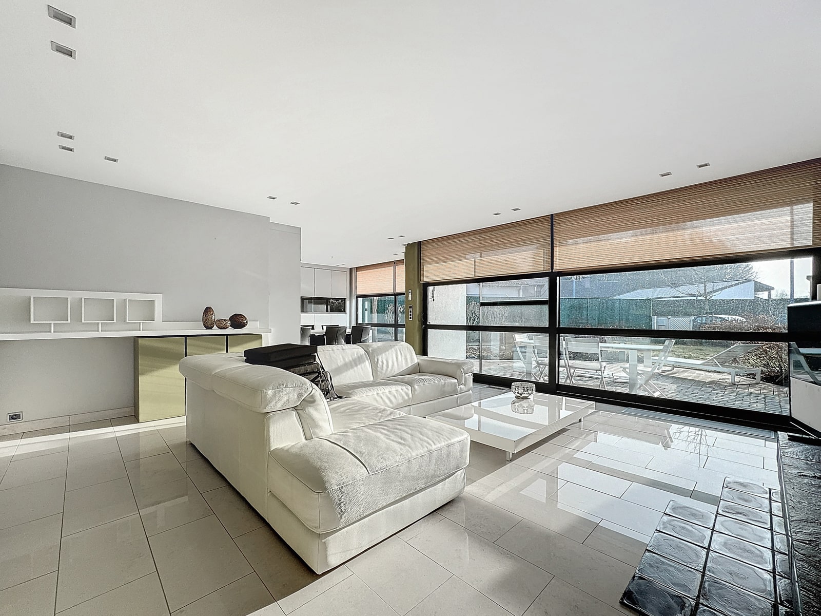 whise photo 5 A modern, sunlit living room with a white leather sectional, a glass coffee table, and large windows with wooden blinds overlooking a patio.