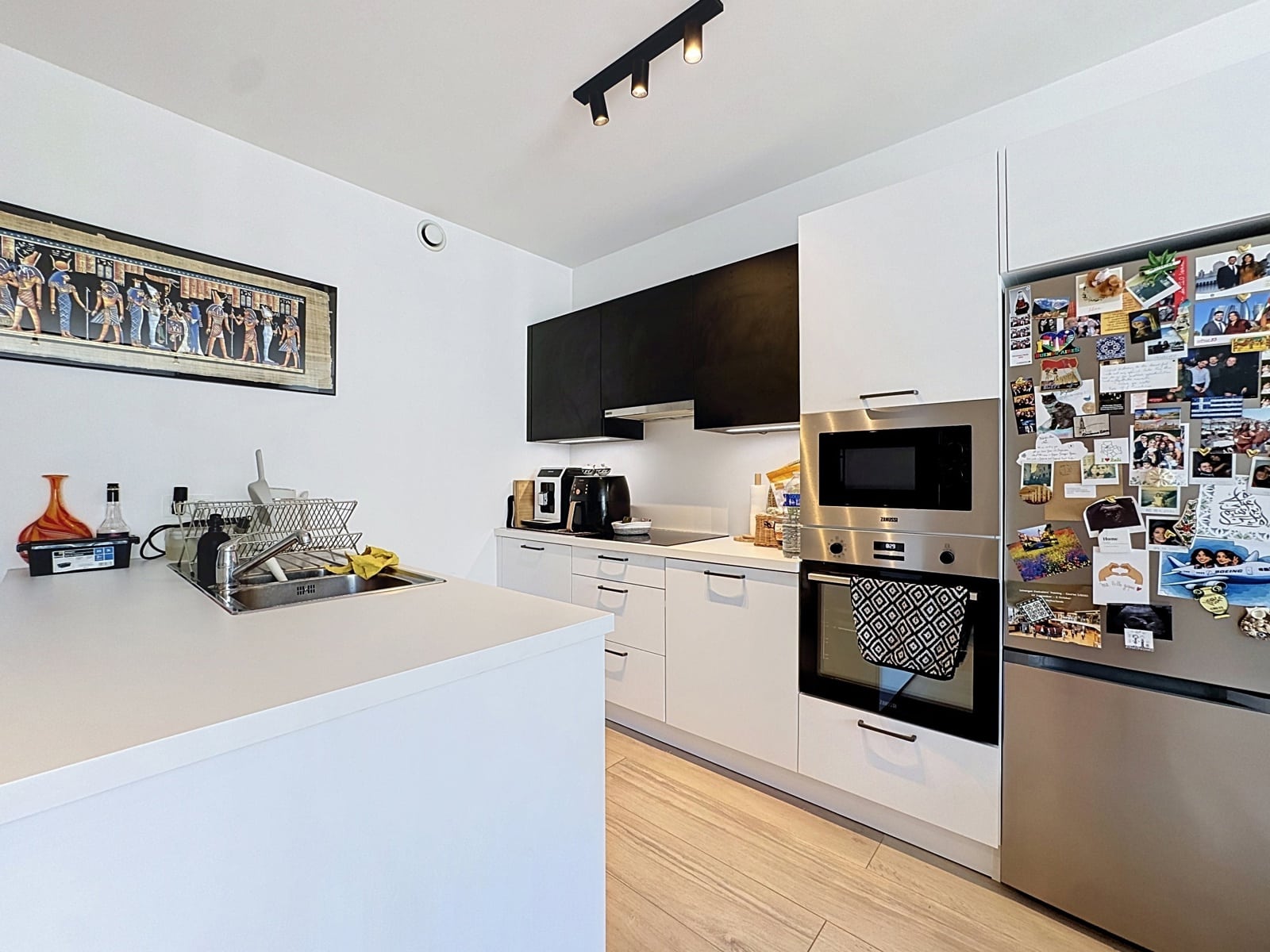 Cuisine Moderne Noir et Blanc – You Real Estate Agency Modern white kitchen with stainless fridge covered in magnets, oven, microwave, and a sink on a white island.