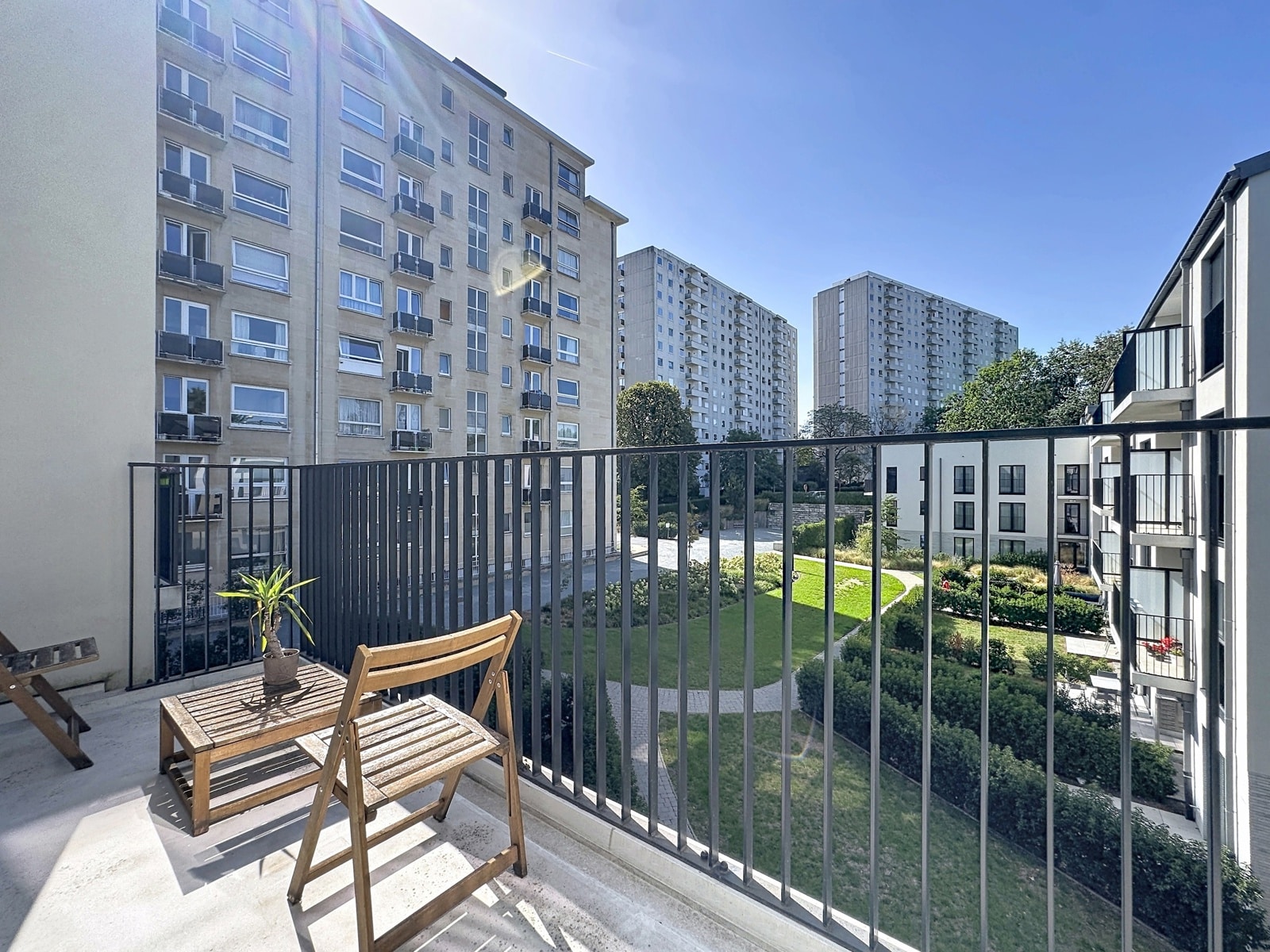 Balcon Ensoleillé avec Vue sur Cour – You Real Estate Agency Terrasse d’appartement ensoleillée avec vue sur cour paysagée et résidences modernes.