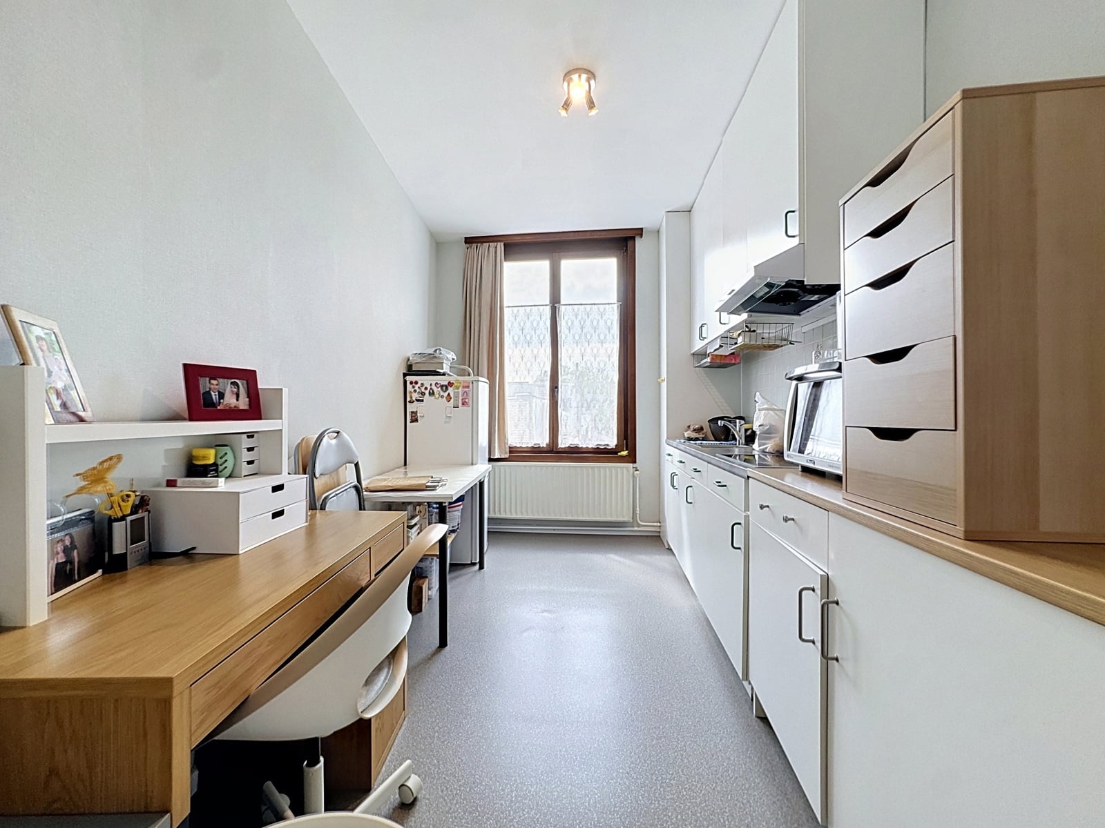 whise photo 11 Bright galley kitchen with white cabinets on the right, a wooden table and chair on the left, and a small fridge by the window at the end.