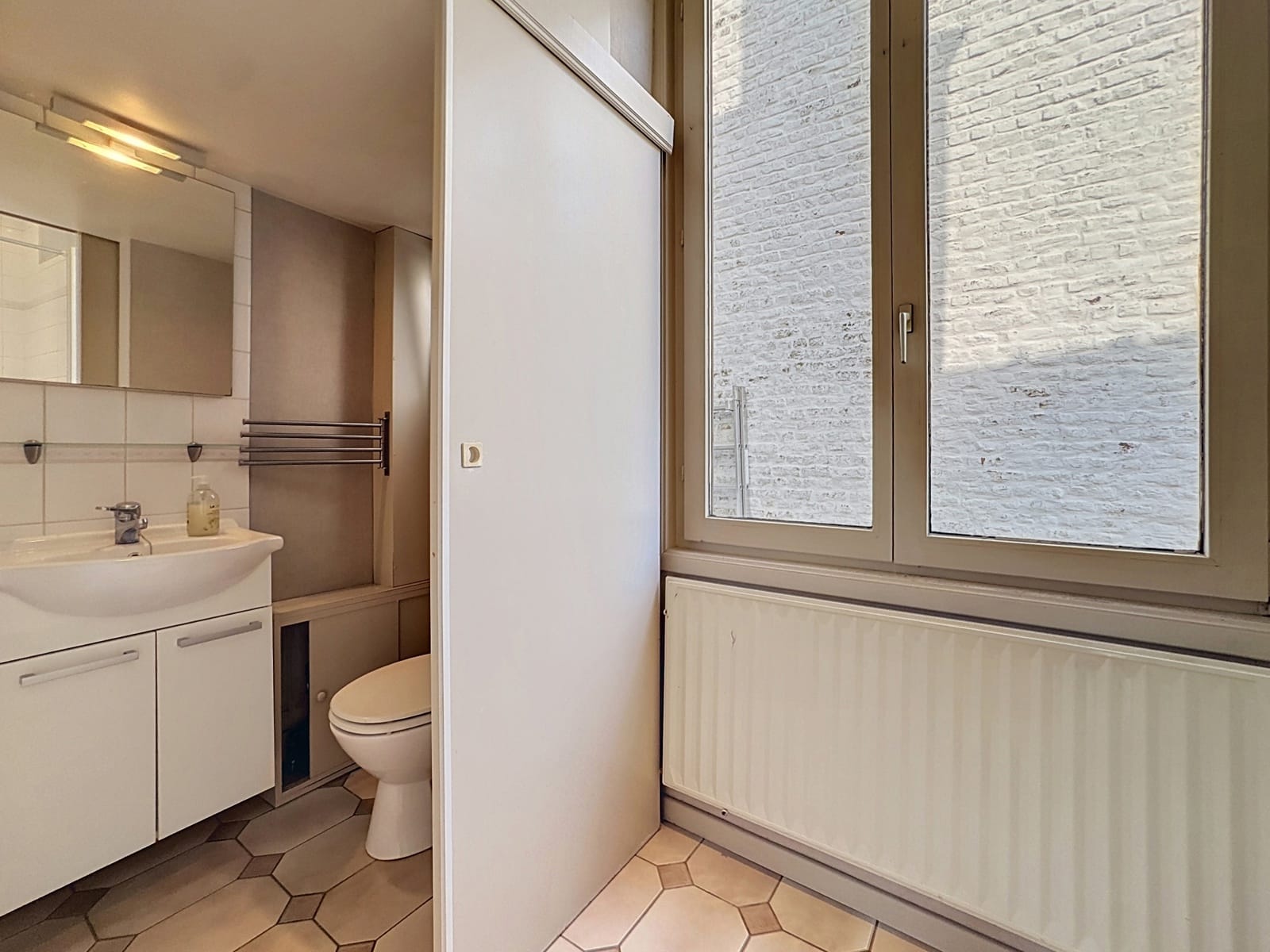 whise photo 8 Small bathroom with white vanity, sink, mirror, and towel rack next to a toilet, divided by a sliding door, large window showing a brick wall outside, and a radiator beneath the window.