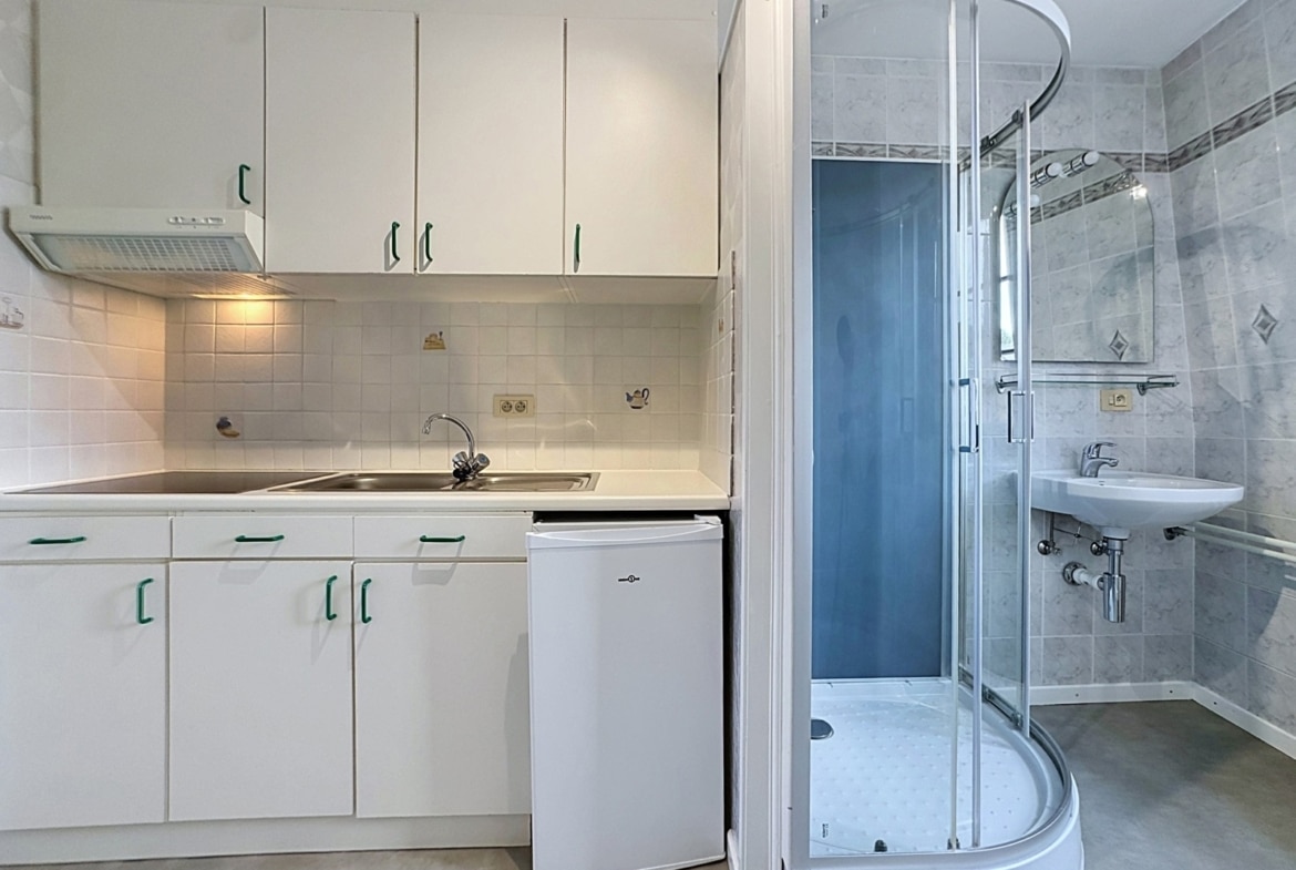 White kitchenette with upper and lower white cabinets, sink, stovetop, and a small fridge; next to a curved glass shower enclosure in a tiled bathroom area