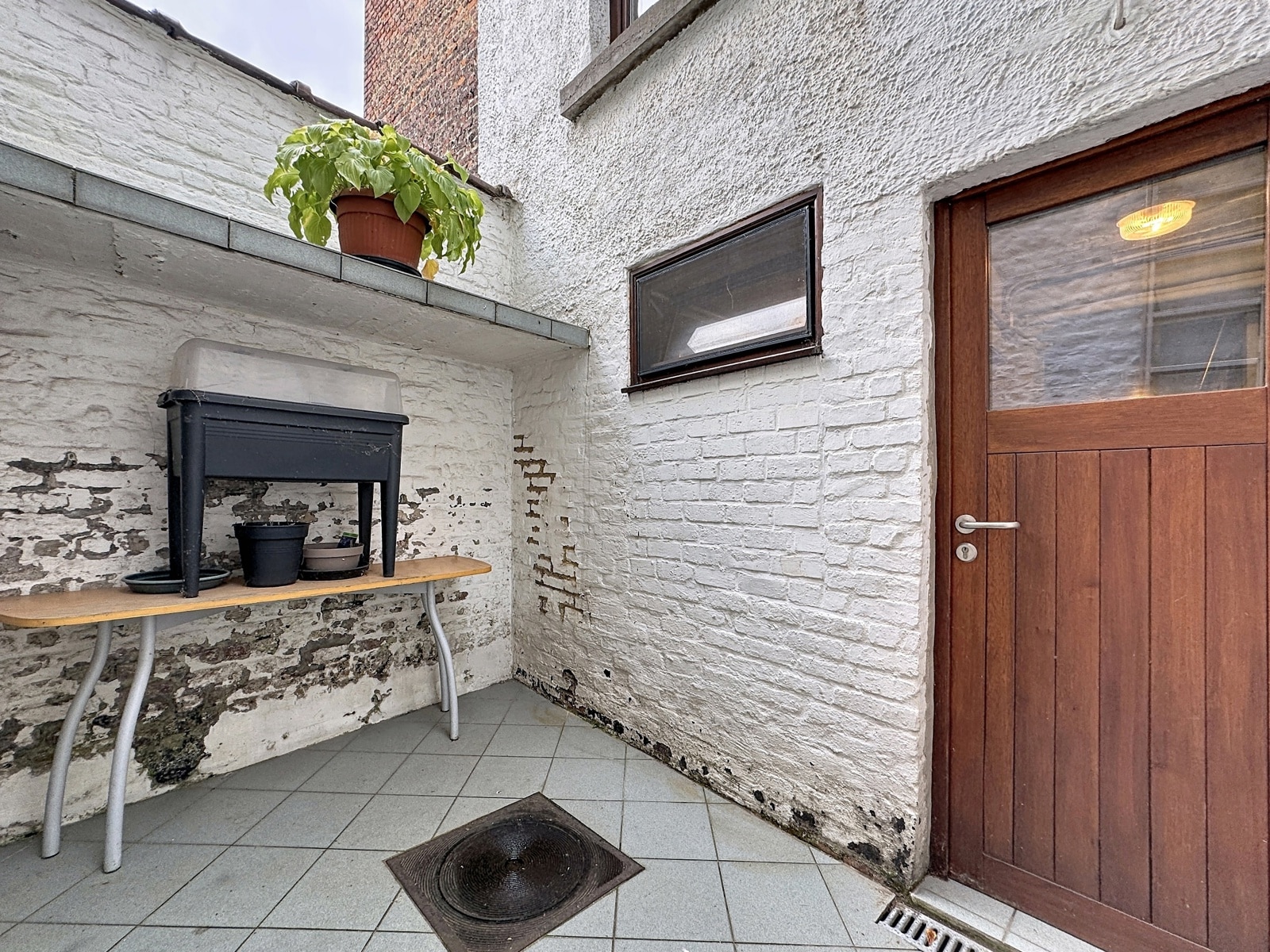 whise photo 28 Small outdoor courtyard with white brick walls, a wooden door, and a potted plant on a ledge above a table holding pots and a black tub.