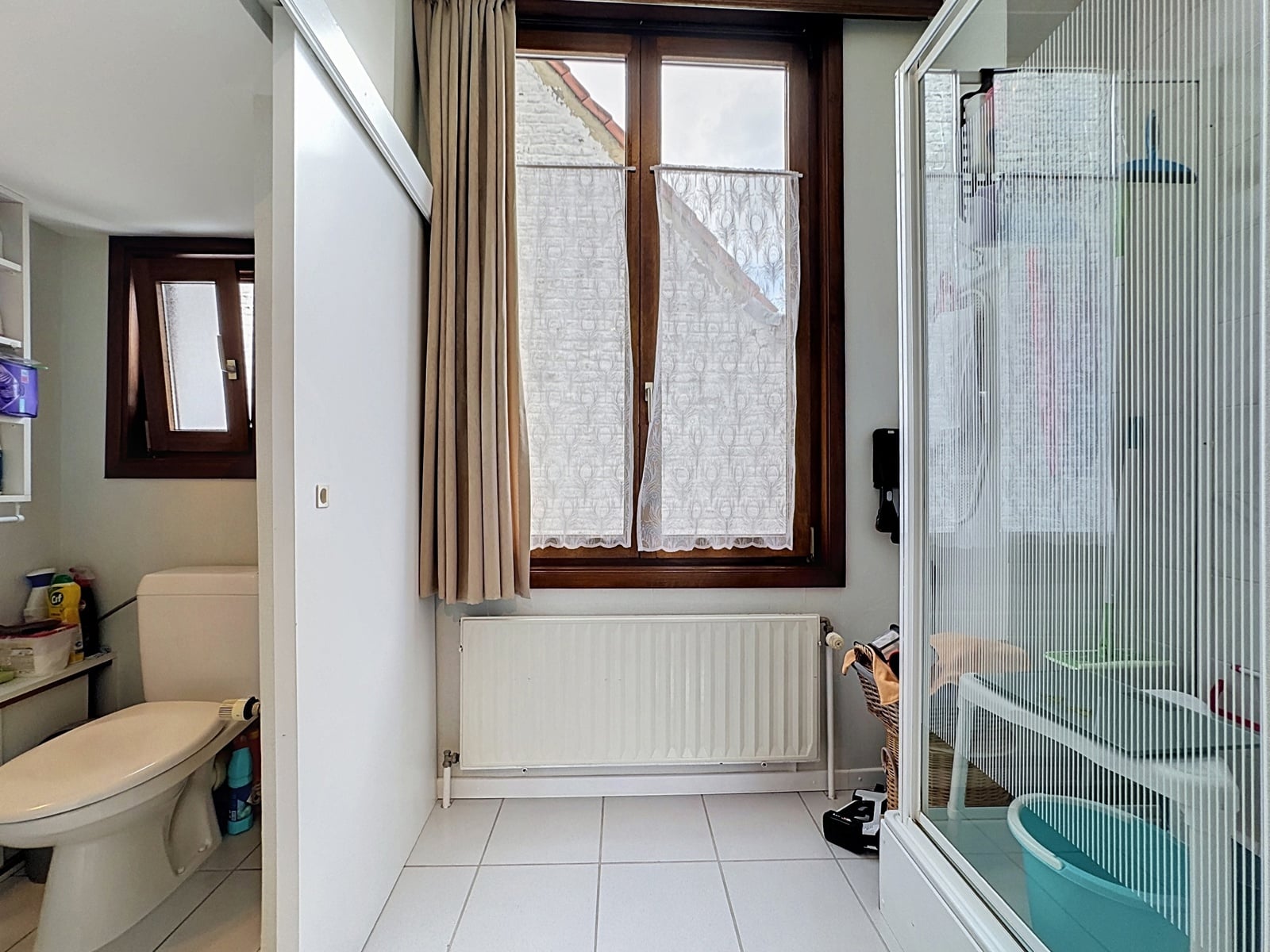 whise photo 16 Bathroom scene with a white toilet on the left, a tall white cabinet, and a wooden-framed window with lace curtains above a radiator in the center. A glass shower enclosure is on the right.