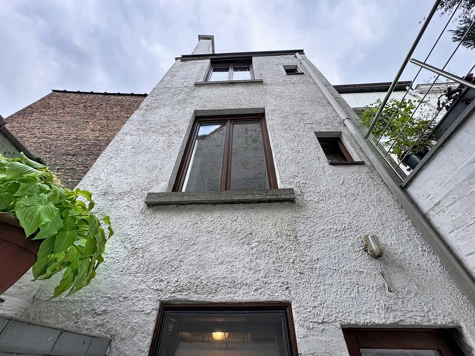 whise photo 30 White stucco townhouse facade seen from ground level, with a tall central window and a brick roof to the left under a cloudy sky.